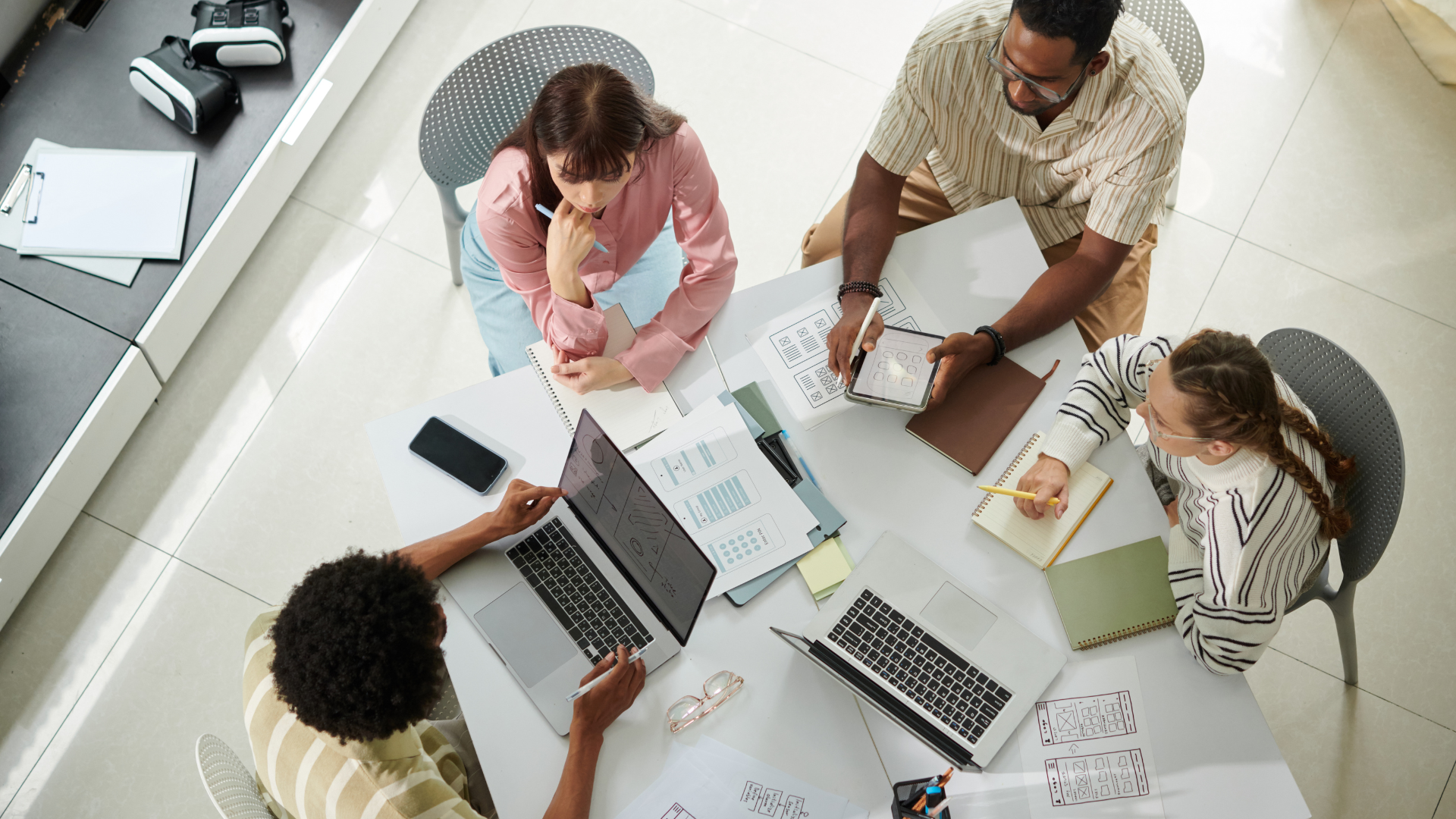 A diverse group of five people gathered around a table working on laptops, tablets, notebooks, and documents in a modern office.