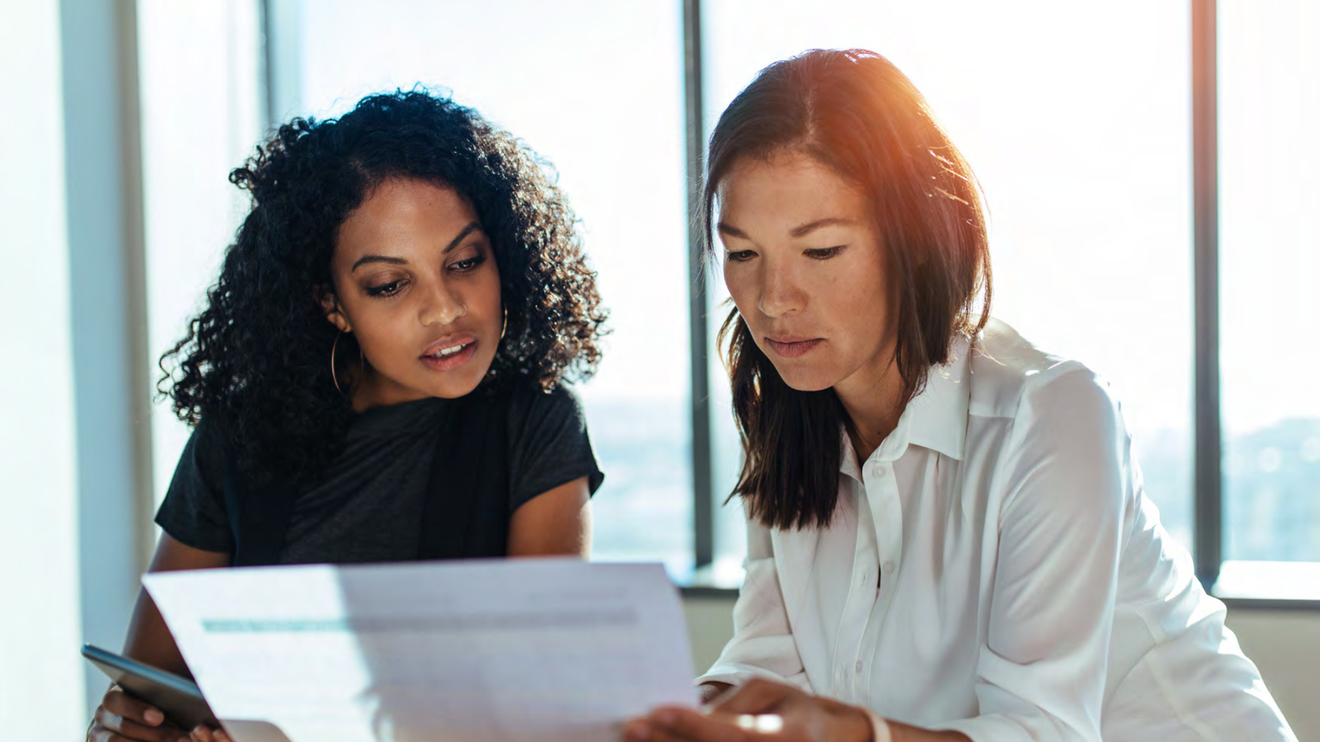 Two women looking at a document or paper in an office with large windows letting in natural light.