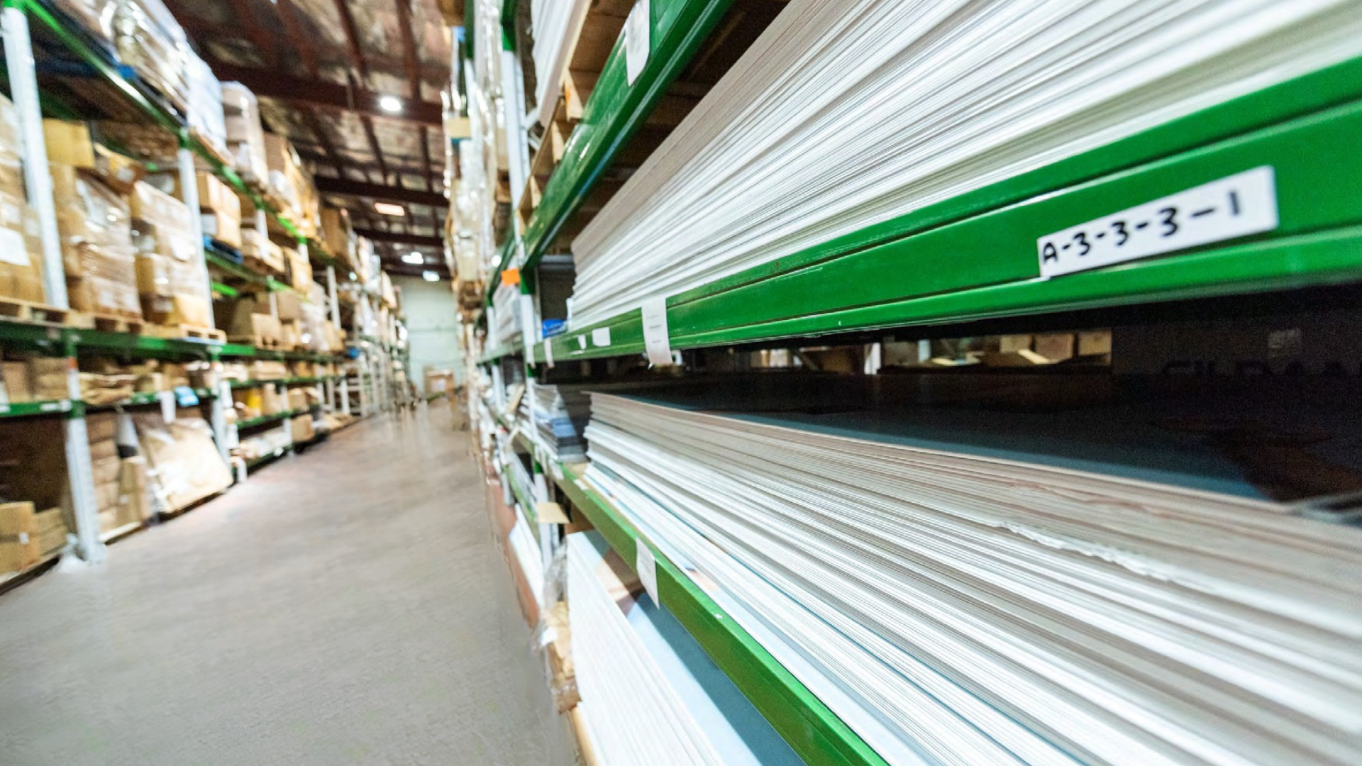 A warehouse aisle with shelves filled with paper rolls and boxes, viewed from ground level looking down the aisle.