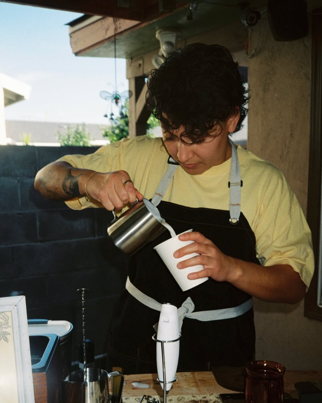 Person pouring milk into a cup under a mounted blender on a wooden table inside a kitchen or cafe.