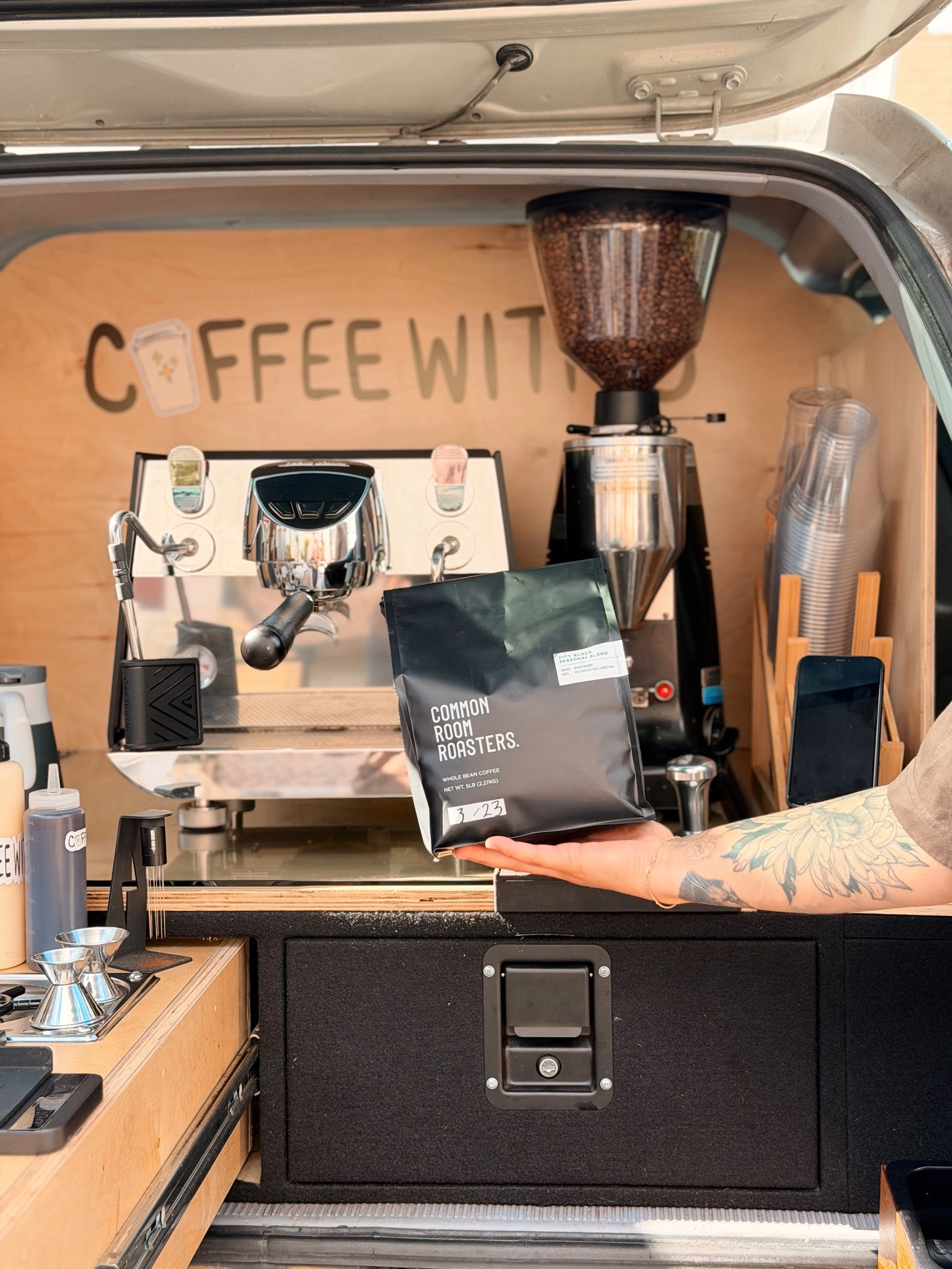 Inside a coffee truck, a person with tattoos on their arm holds a bag of coffee labeled 'Common Room Roasters.' The background displays a coffee machine, grinder, and cups, with a sign that reads 'Coffee Witt' on the wall.