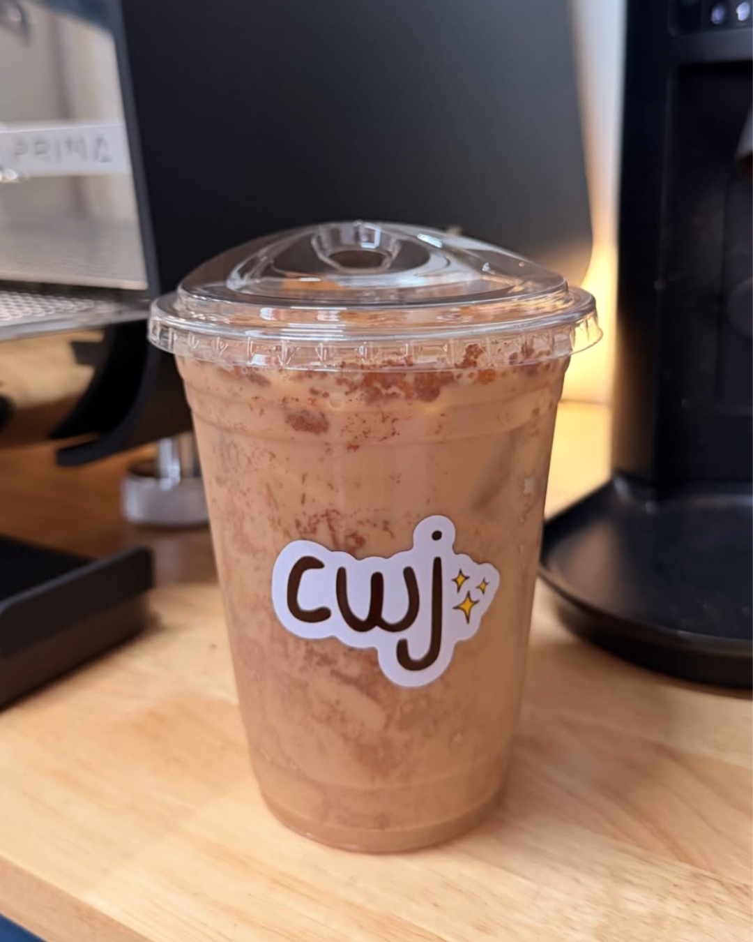 A cold beverage in a plastic cup with a clear lid, placed on a wooden surface, with a sticker that says 'wow' decorated with stars, near a coffee machine.