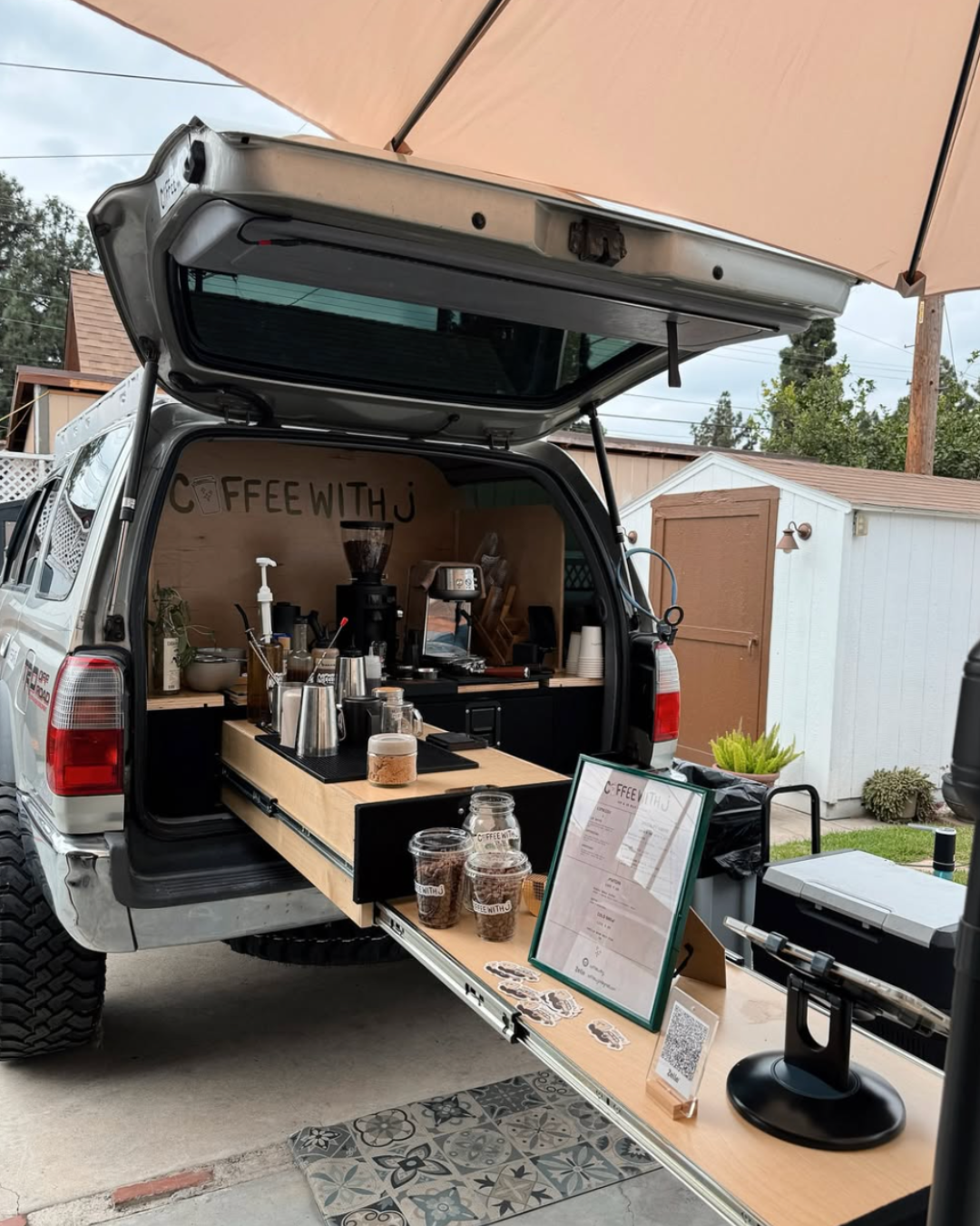 A mobile coffee stand set up inside an open SUV with a folding wooden counter, coffee equipment, cups, and a menu display, under an outdoor umbrella.