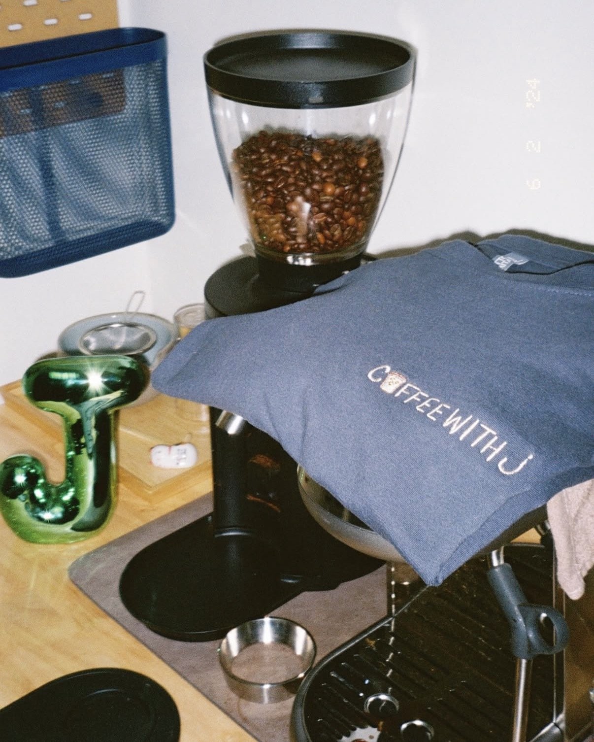 A coffee grinder filled with coffee beans on a countertop next to a blue dishcloth with the logo 'COFFEEWITHJ.' A green decorative 'J' and various coffee accessories are also visible.