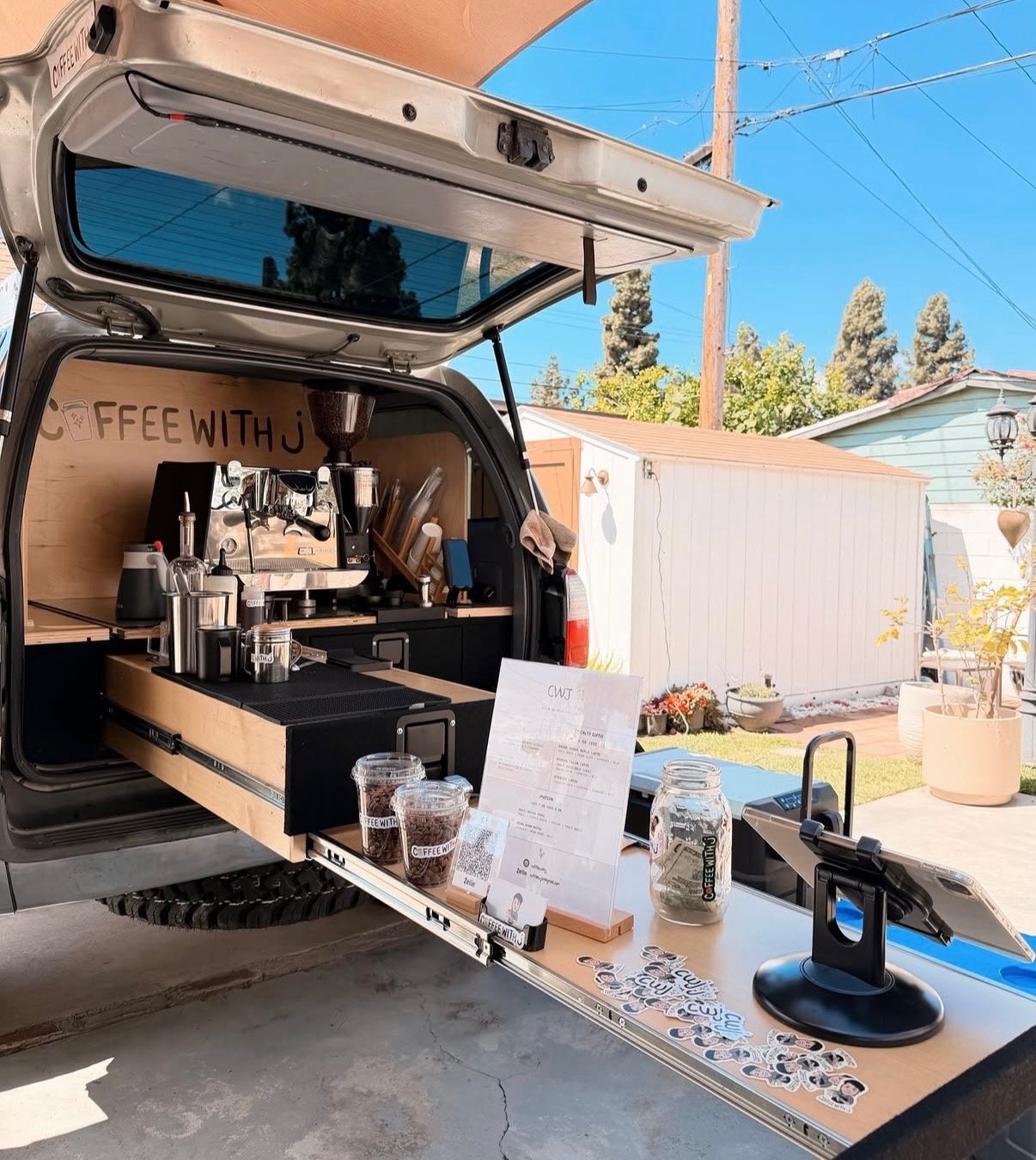 A black mobile coffee truck with an open hatch showing a coffee brewing station inside. The truck's side counter extends outward, displaying menu, stickers, and jars of ingredients, with a tablet on a stand for orders. Outside, bright sunny weather, trees, and a backyard with a shed, plants, and outdoor chairs are visible.