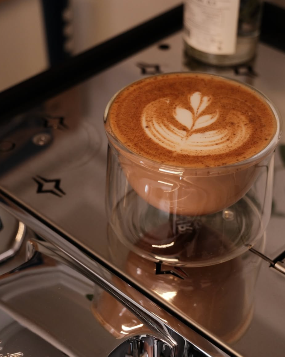 A glass cup filled with hot coffee with latte art in the shape of a leaf, placed on a silver espresso machine.