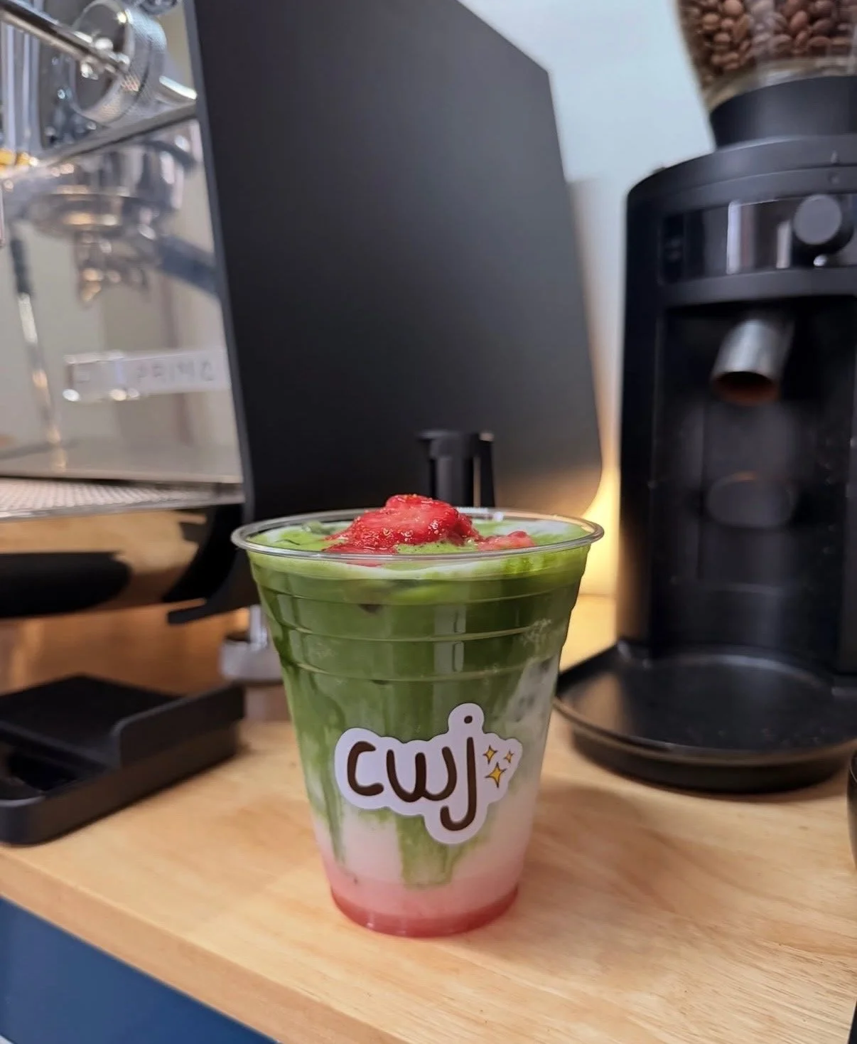 A layered green and pink drink with a red topping in a clear plastic cup, placed on a wooden surface.