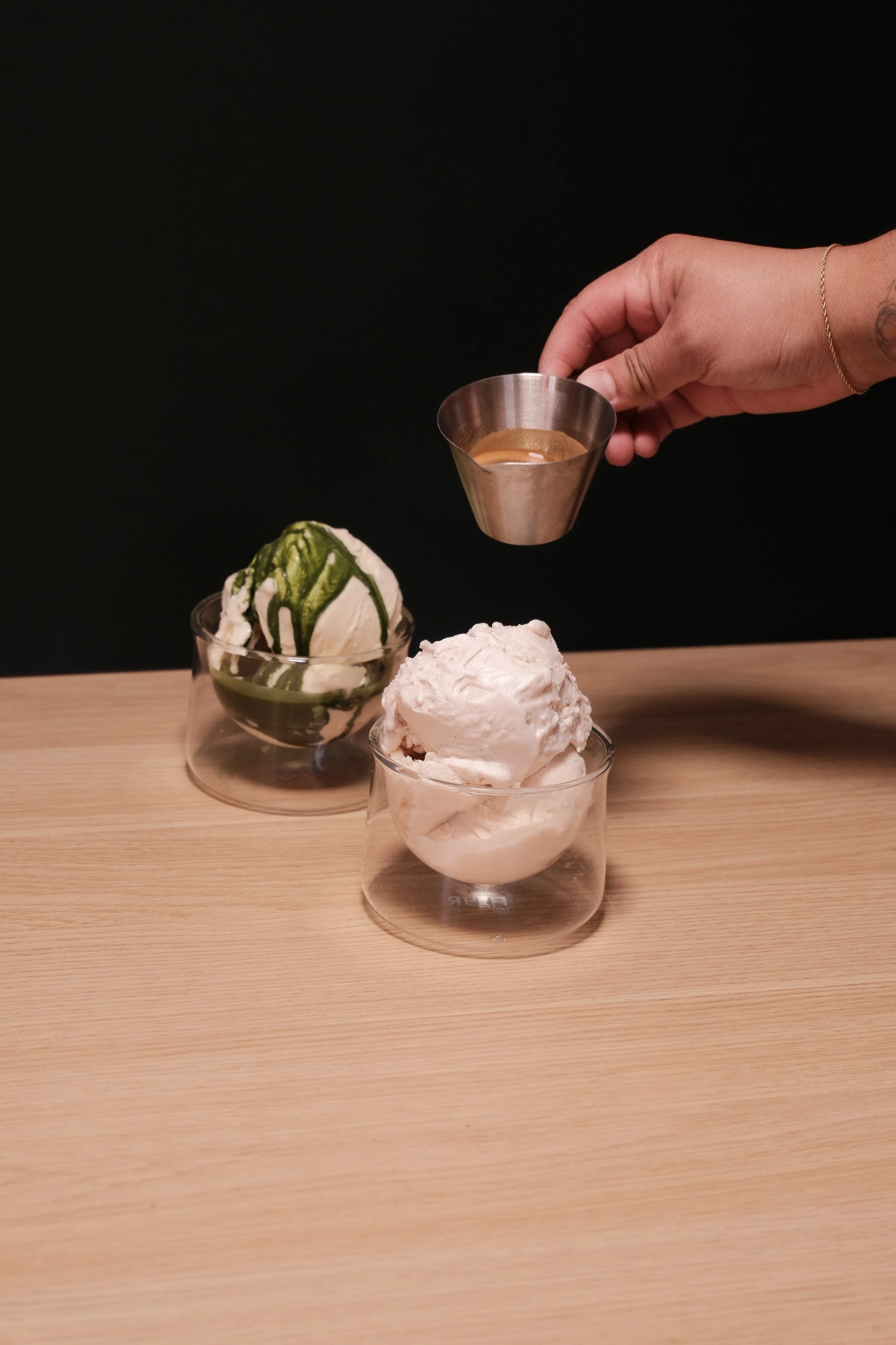 Hand holding a metal espresso shot glass over an ice cream dessert with green sauce on top