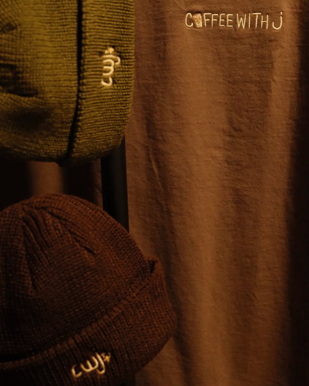 Close-up of clothing with embroidered text: "COFFEE WITH J" on a brown fabric, an olive green textured cap with embroidered number "33", and a maroon checkered cap.