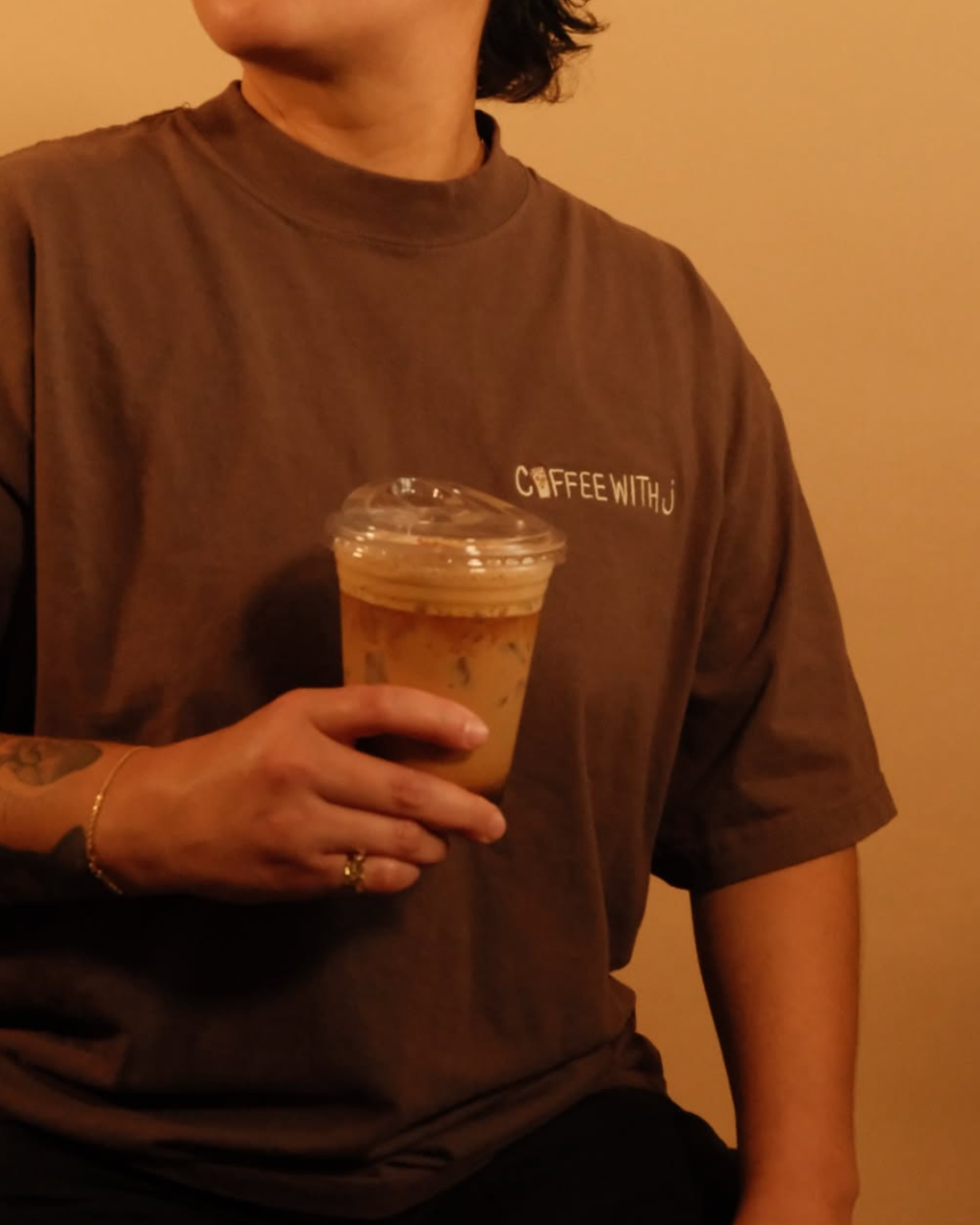 Person holding a plastic cup of iced coffee with a dome lid, wearing a brown t-shirt with the logo 'COFFEE WITH J' in white text.
