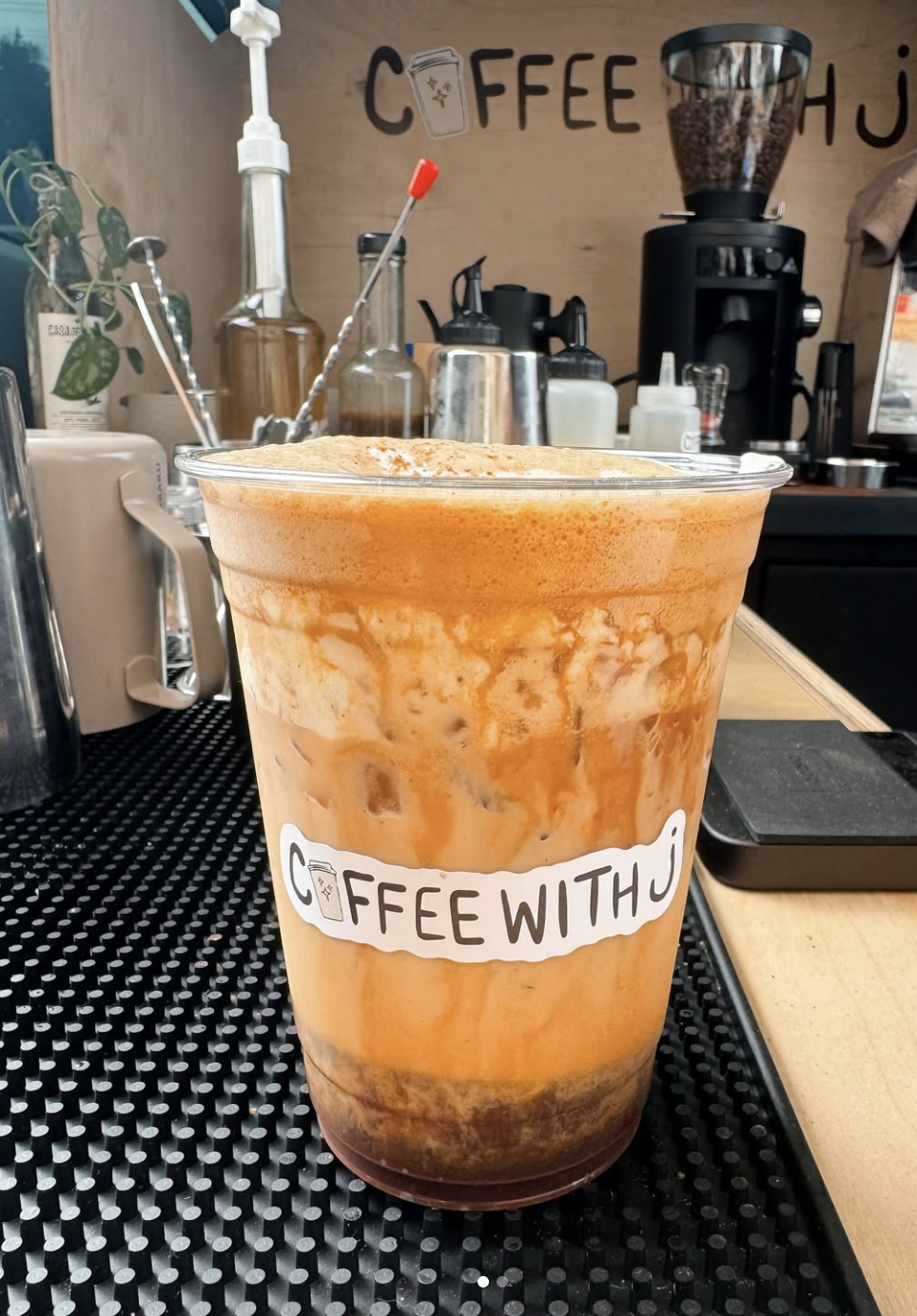 A caramel frappuccino with whipped cream in a clear plastic cup labeled 'Coffee With U' on a coffee bar counter.