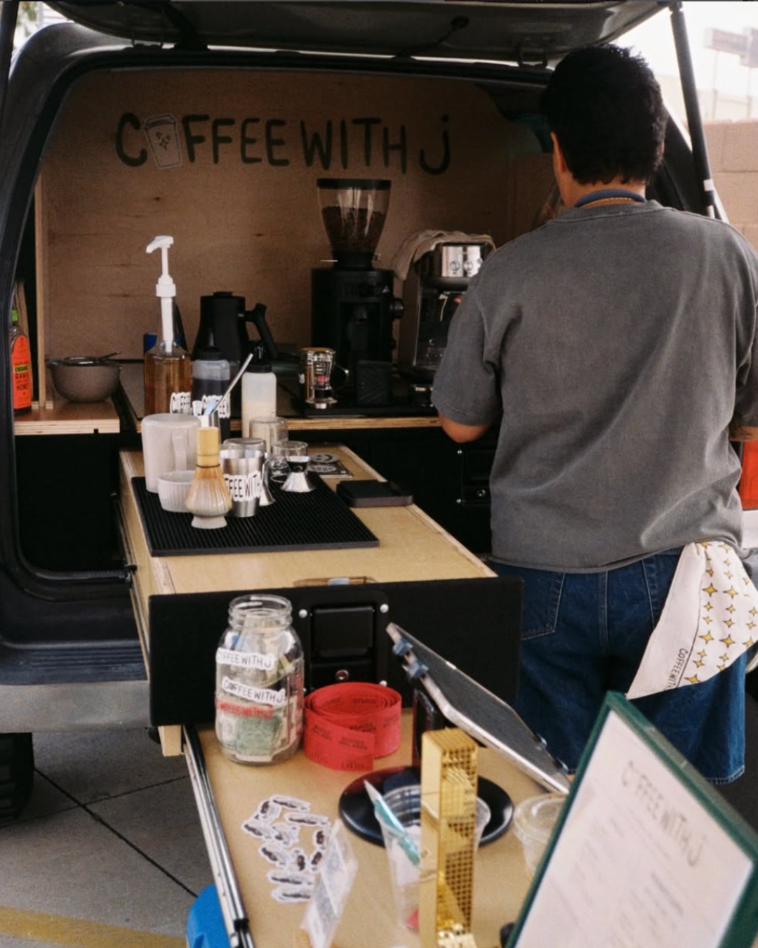 A person making coffee at a mobile coffee stand with a sign that says 'Coffee with J' on the wall behindd the counter. The stand has various coffee-making equipment and supplies.
