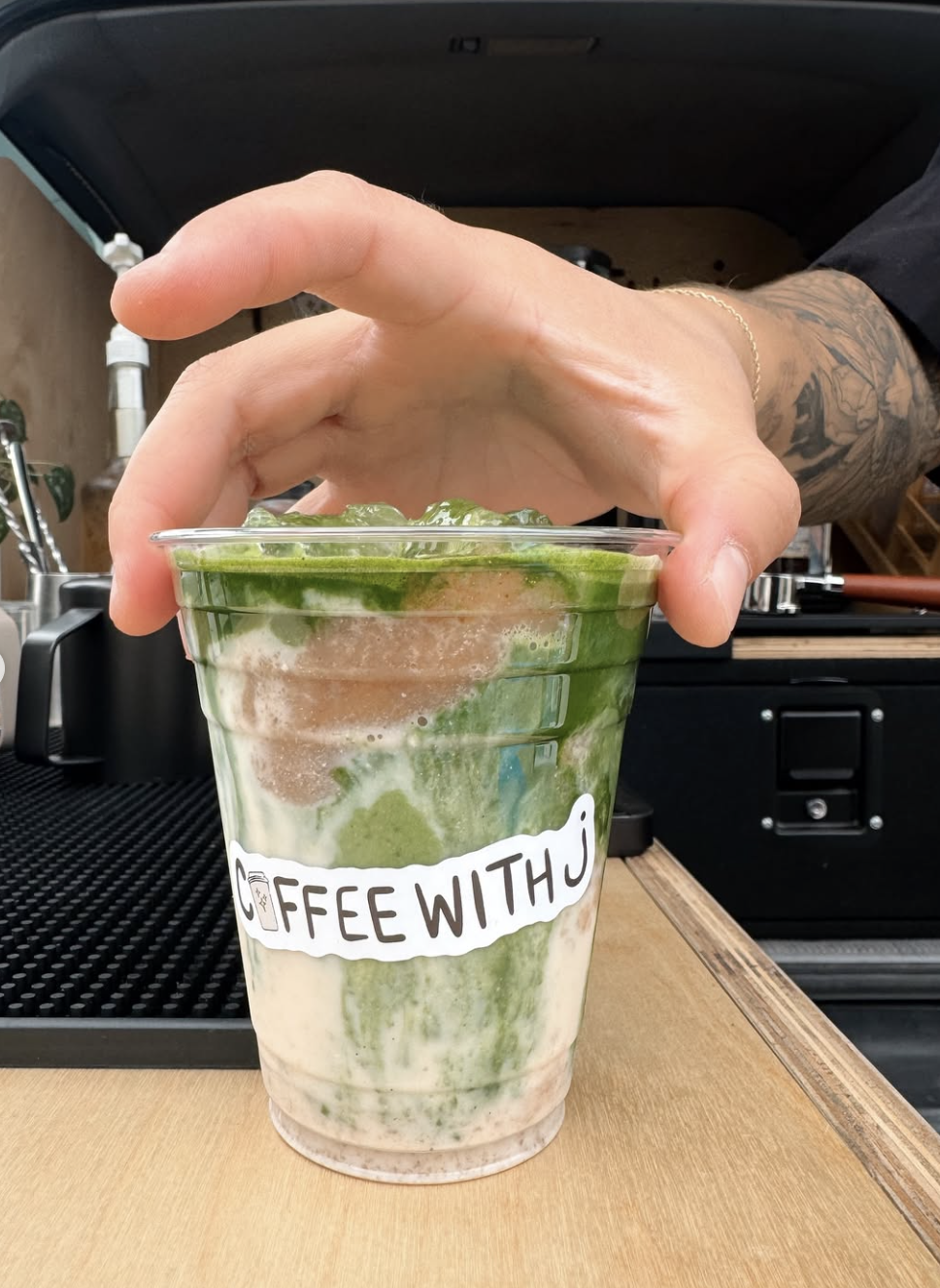 A hand with tattoos holds a clear plastic cup filled with a layered green and white latte, topped with ice, on a wooden surface in front of a black espresso machine. The cup has a sticker that reads 'COFFEE WITH J'.