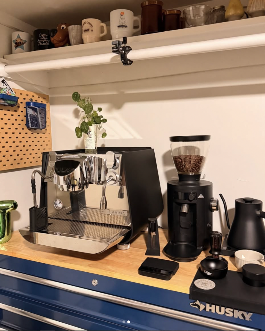 Coffee setup on a wooden countertop with an espresso machine, a coffee grinder filled with beans, and various cups and accessories. A small plant in a glass container is also present.