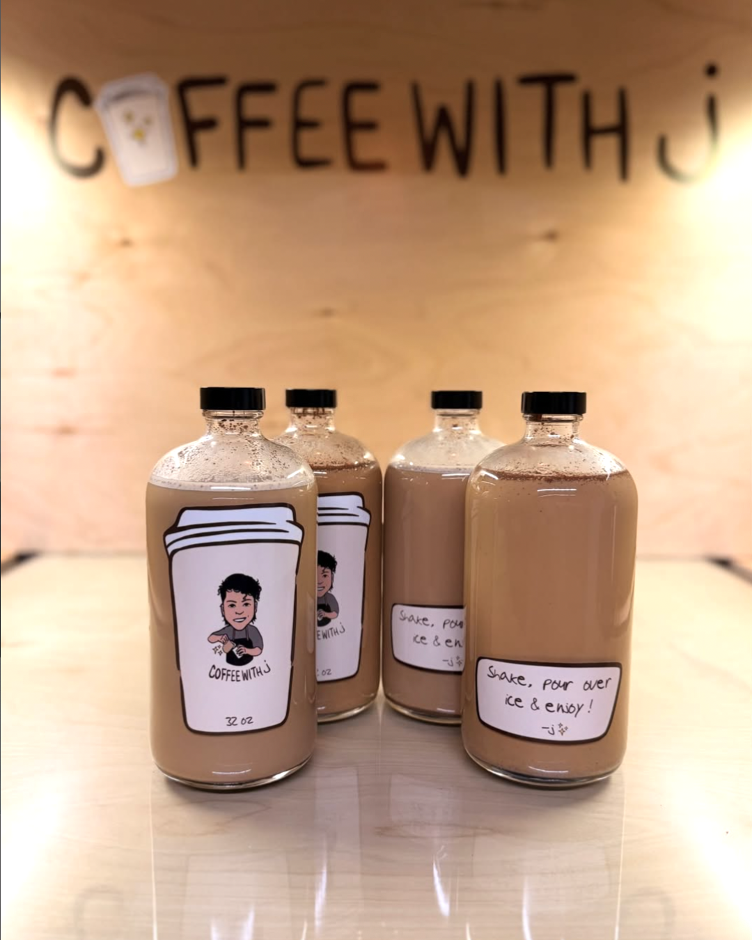 Four bottles of a drink labeled 'CoffeeWithJ' on a wooden surface with a light wooden wall in the background. The bottles have black caps and a cartoon face on the label. The wall in the background has the phrase 'COFFEE WITH J' written on it.