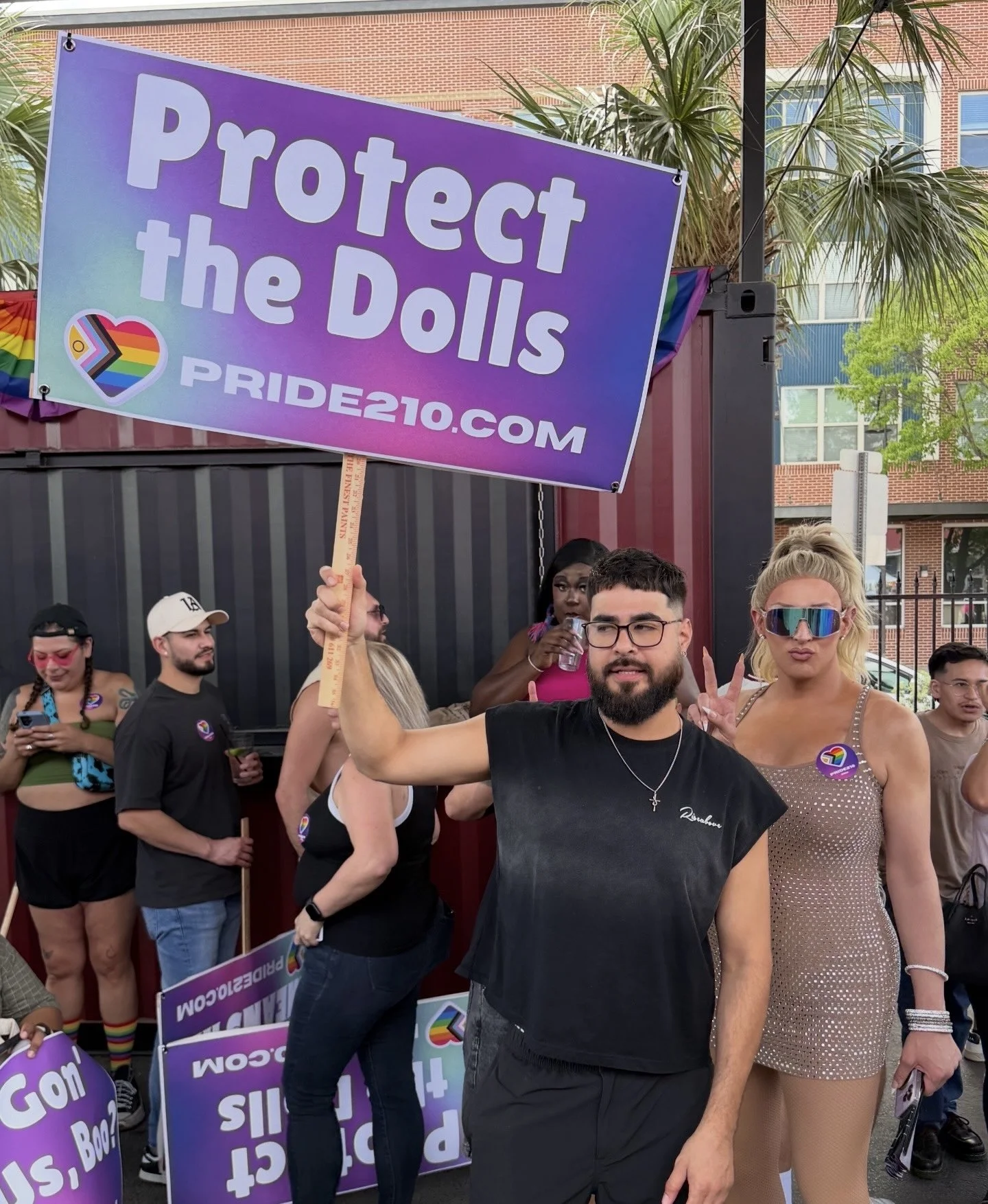 LGBTQ+ community members at a San Antonio Pride event holding a “Protect the Dolls” sign with Pride210.com, representing the Pride210 movement for accountability in San Antonio Pride.