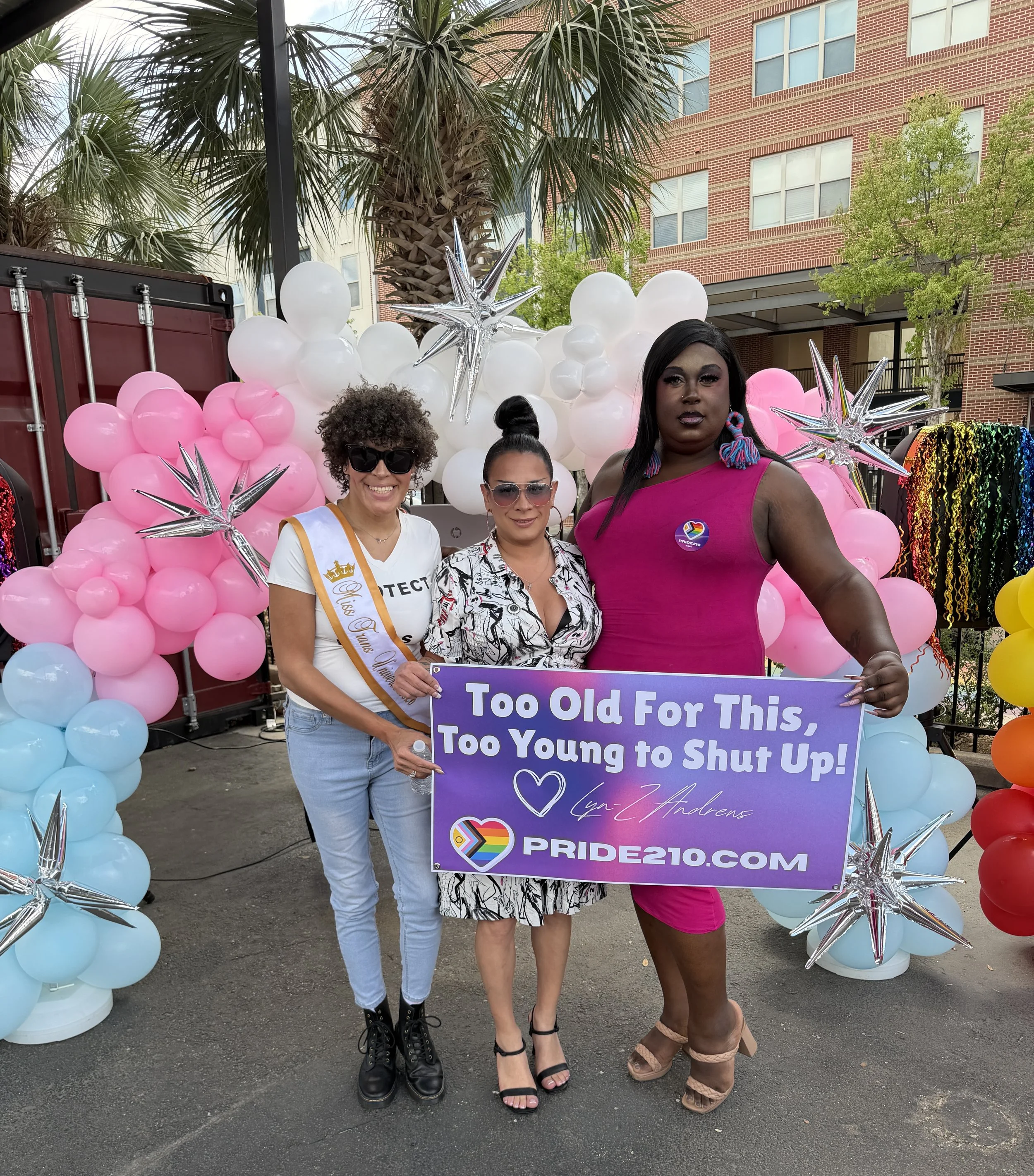 Lyn-Z Andrews, Monique Fontaine-Flores, and Antonia Hall at a San Antonio Pride event holding a Pride210 sign reading “Too Old For This, Too Young to Shut Up!” in front of rainbow balloons.
