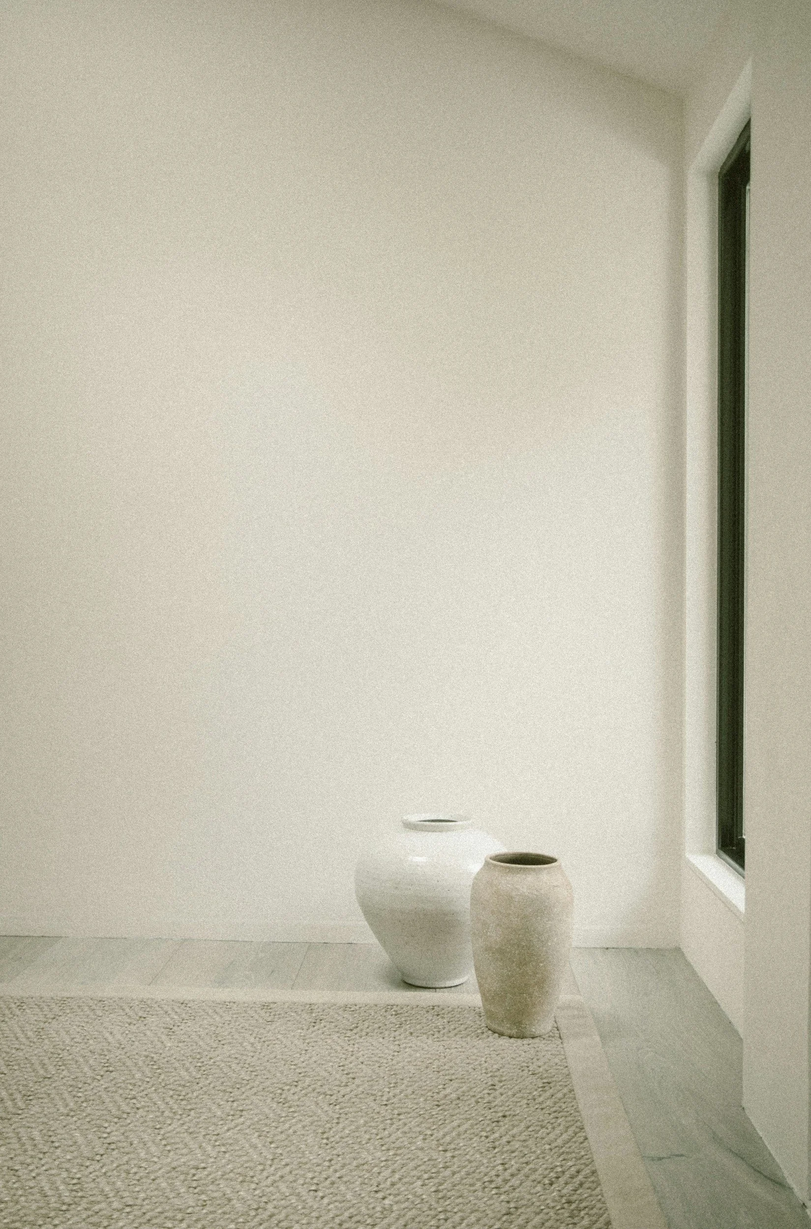 Minimalist interior with a white wall, a window on the right, a beige carpet, and two decorative ceramic vases on the floor.