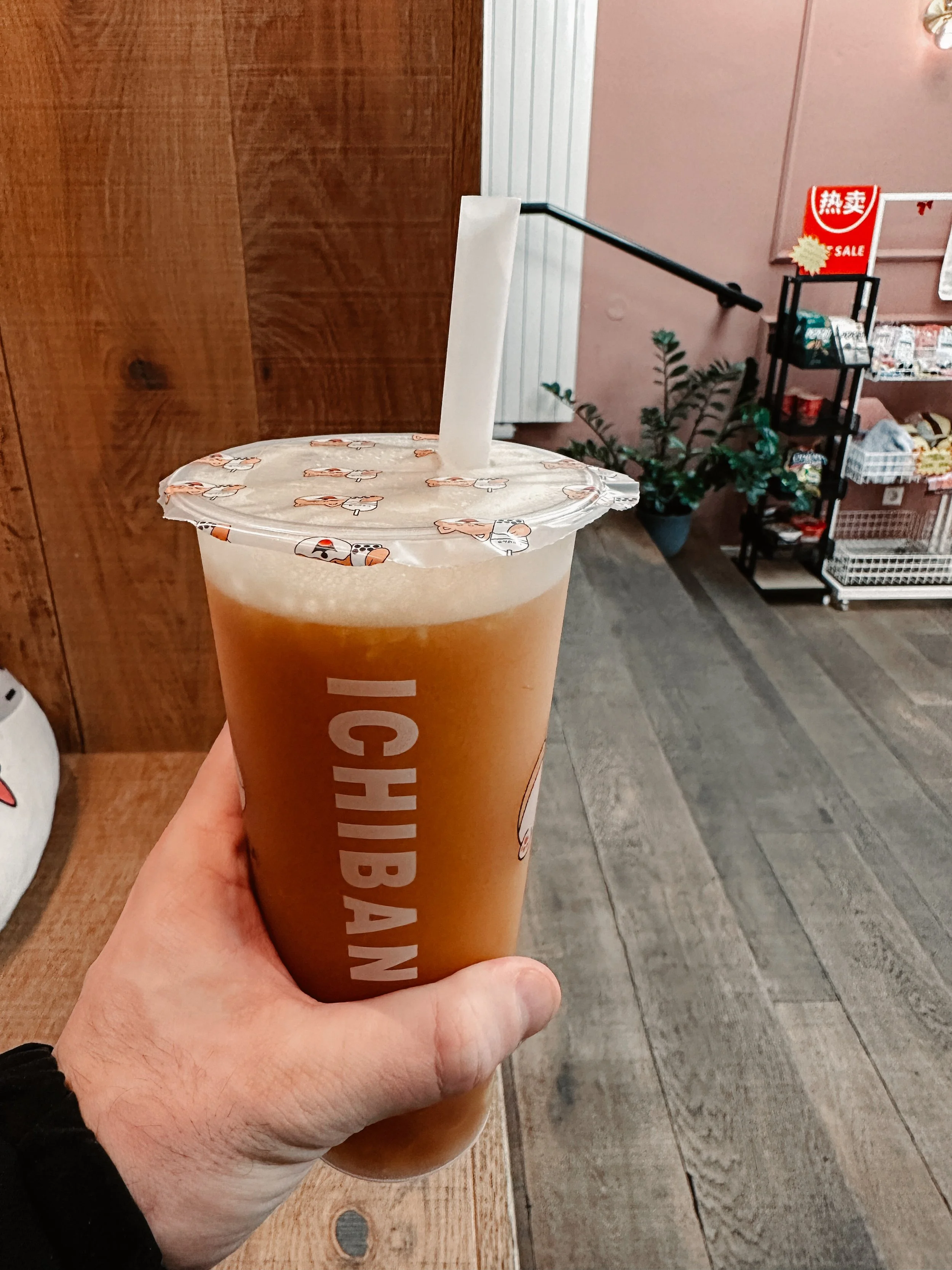 Bottiglia di iced tea con cannuccia, su sfondo di arredi e scaffali di un negozio. Altro a destra, cartello rosso con etichetta di sconto in cinese.