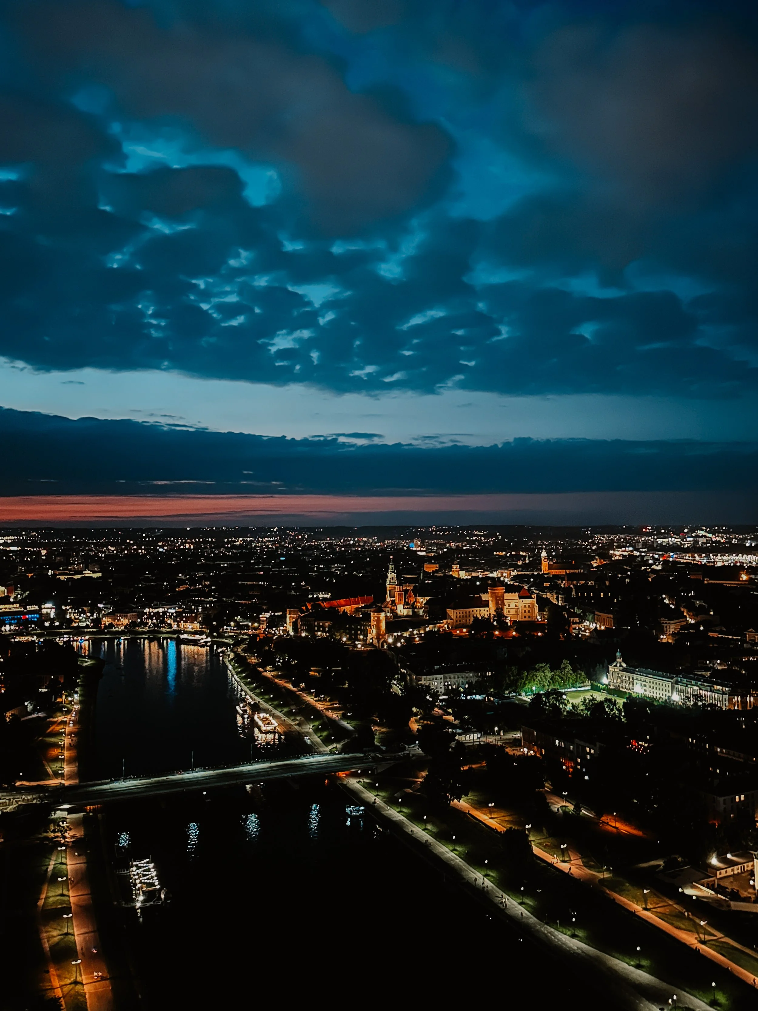 Veduta notturna di una città con edifici storici, un fiume e un ponte, sotto un cielo parzialmente nuvoloso con tonalità di blu e rosa