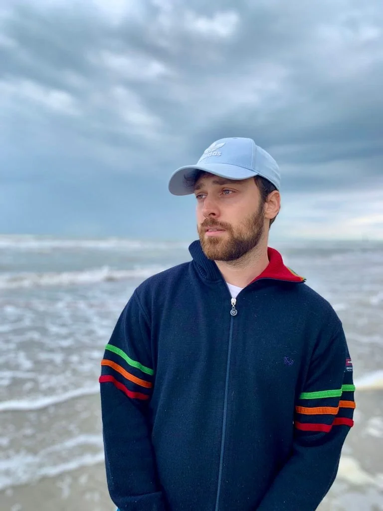 A man with a beard wearing a light gray baseball cap and a dark navy blue jacket with colorful stripes on the sleeves, standing on a beach with waves and overcast sky in the background.