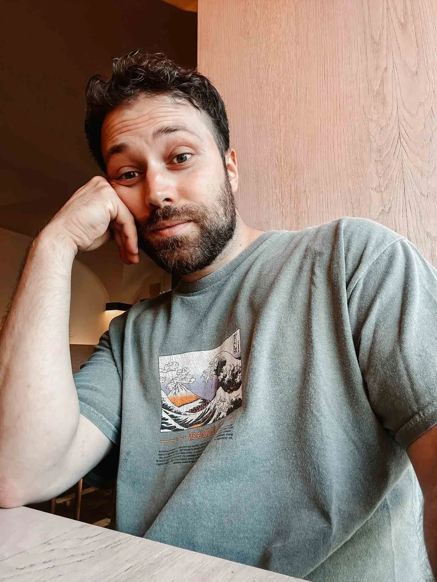 A man with dark hair and a beard sitting at a table, resting his head on his left hand, wearing a grey T-shirt with a graphic design, in a cozy indoor setting.