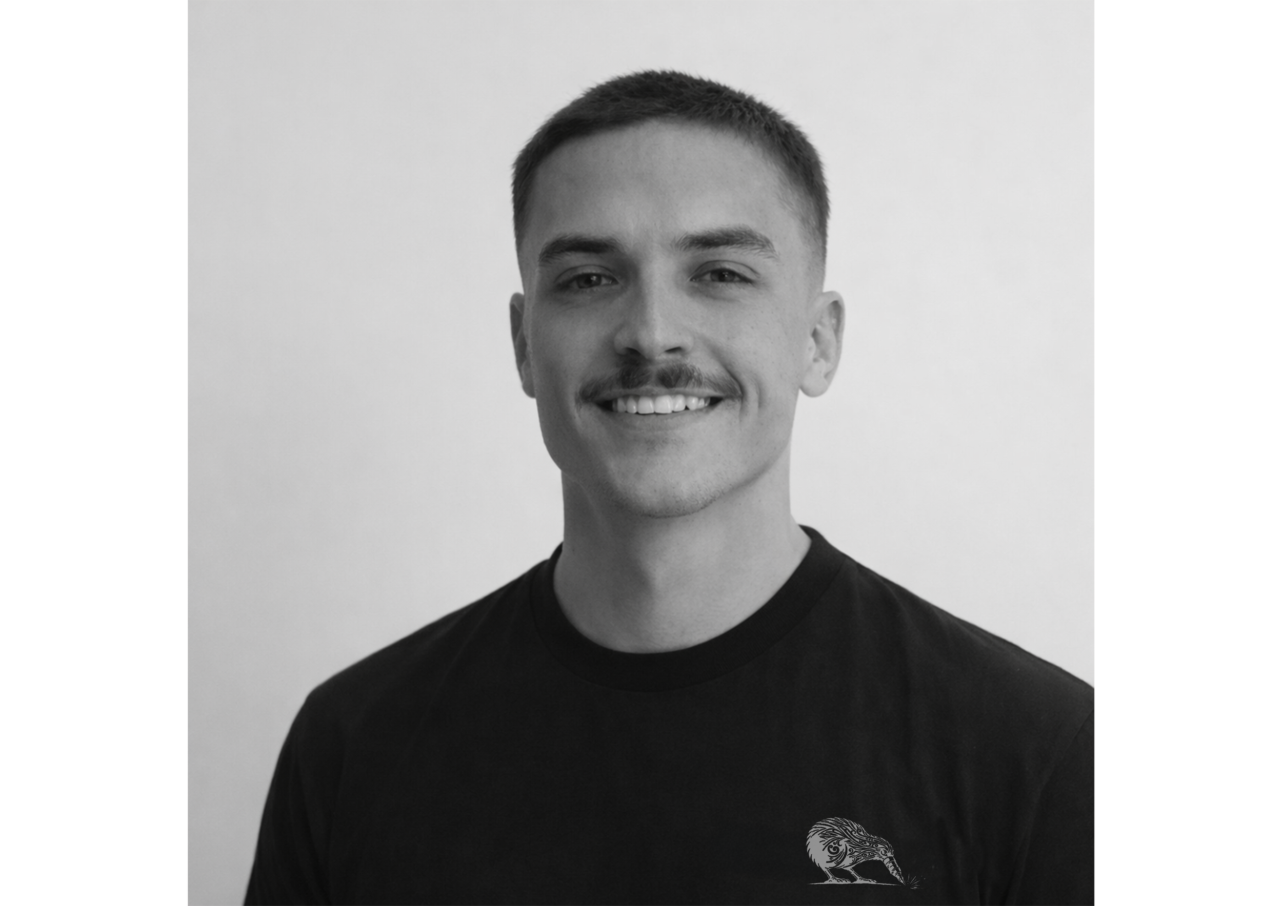 Black and white portrait of a young man with short hair, mustache, smiling, wearing a black T-shirt with a small graphic on the chest, against a plain background.