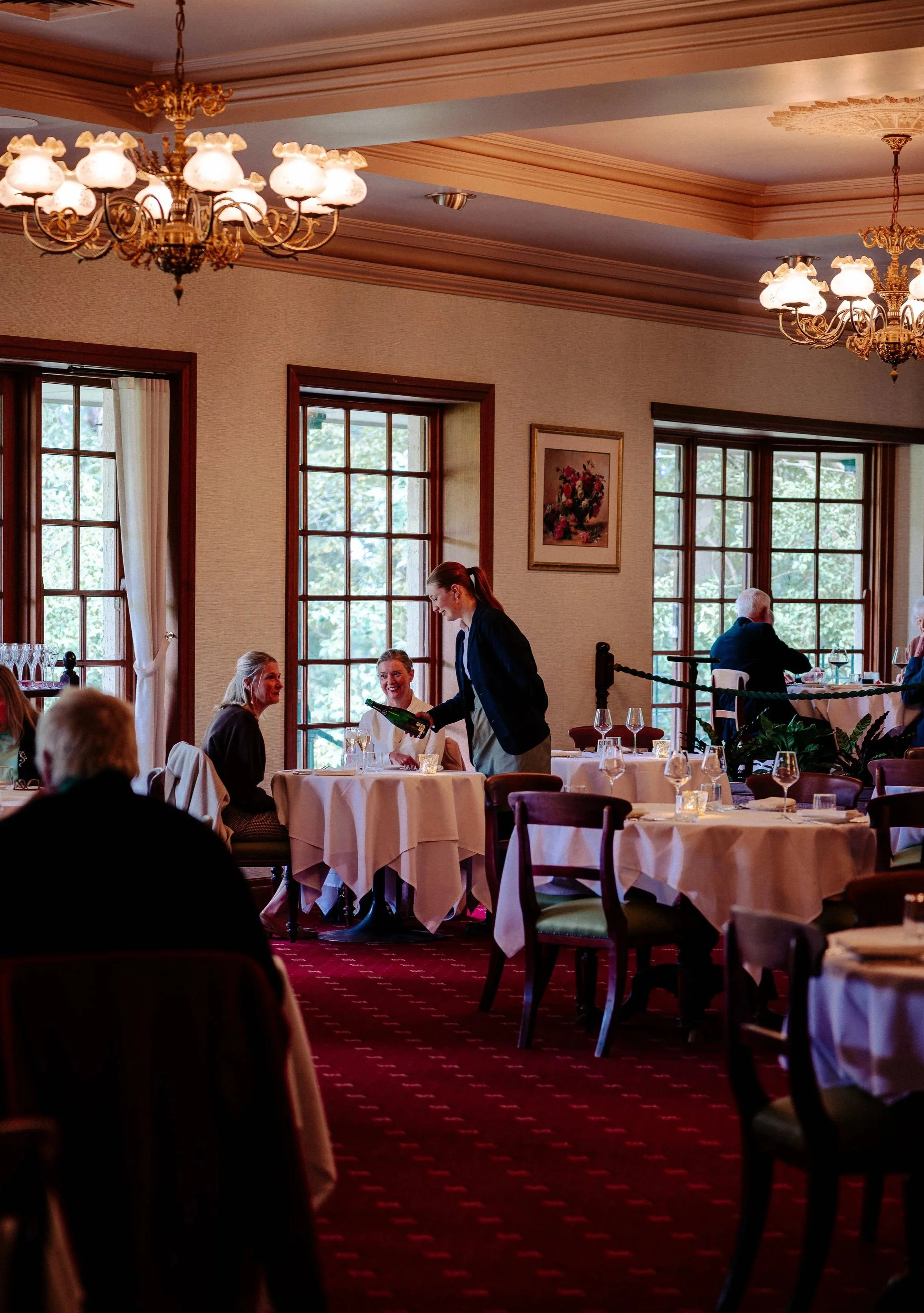 A group of people dining in a restaurant with large windows, chandeliers, and formal table settings, with a server pouring wine at a table.