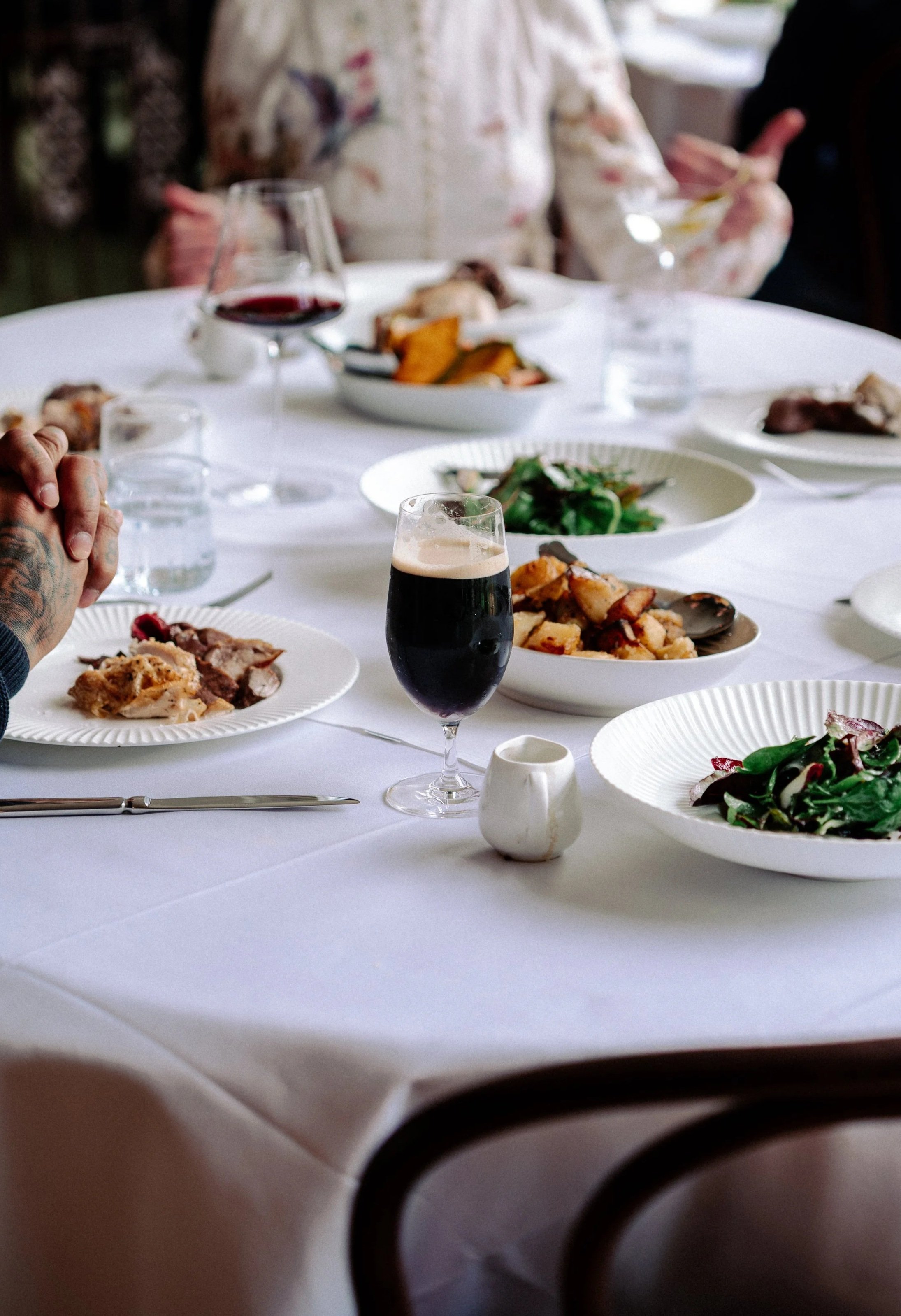A dinner table set with various dishes, glasses of wine and water, with people in the background holding utensils. The focus is on the table and the food.