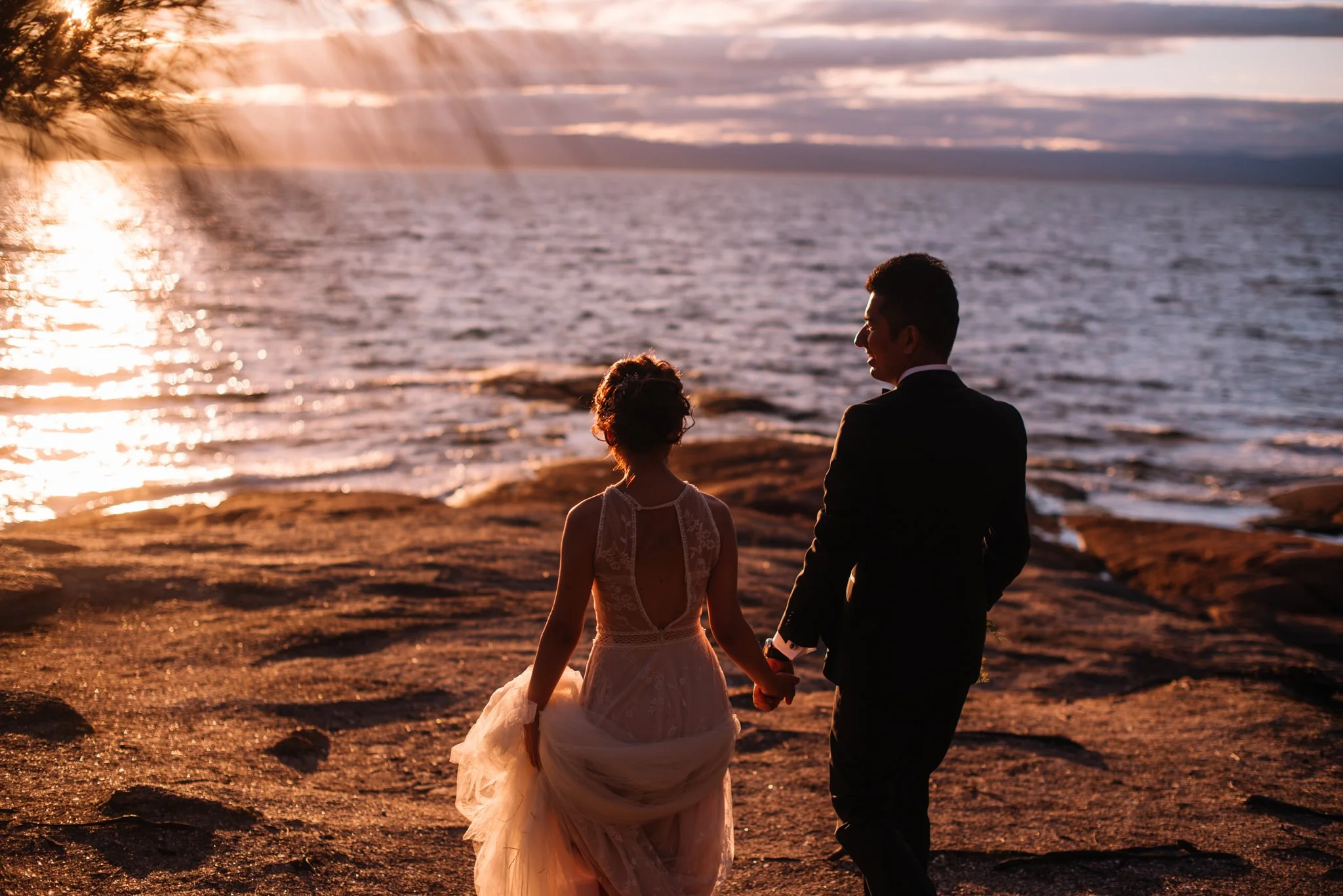 A couple, dressed in wedding attire, holding hands and walking along the beach during sunset.