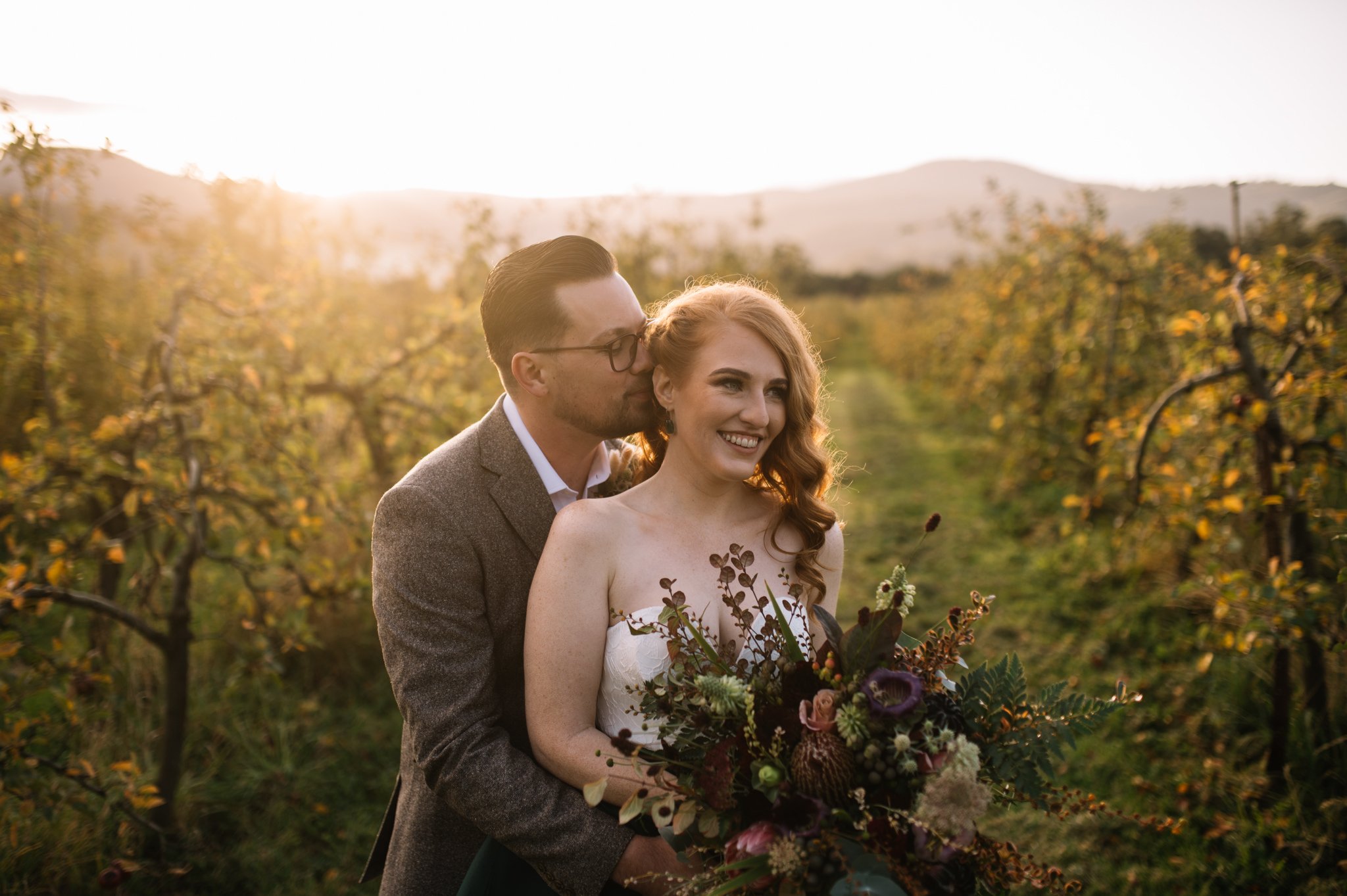 A couple standing in a vineyard during sunset, with the woman holding a bouquet of flowers and the man kissing her on the cheek.