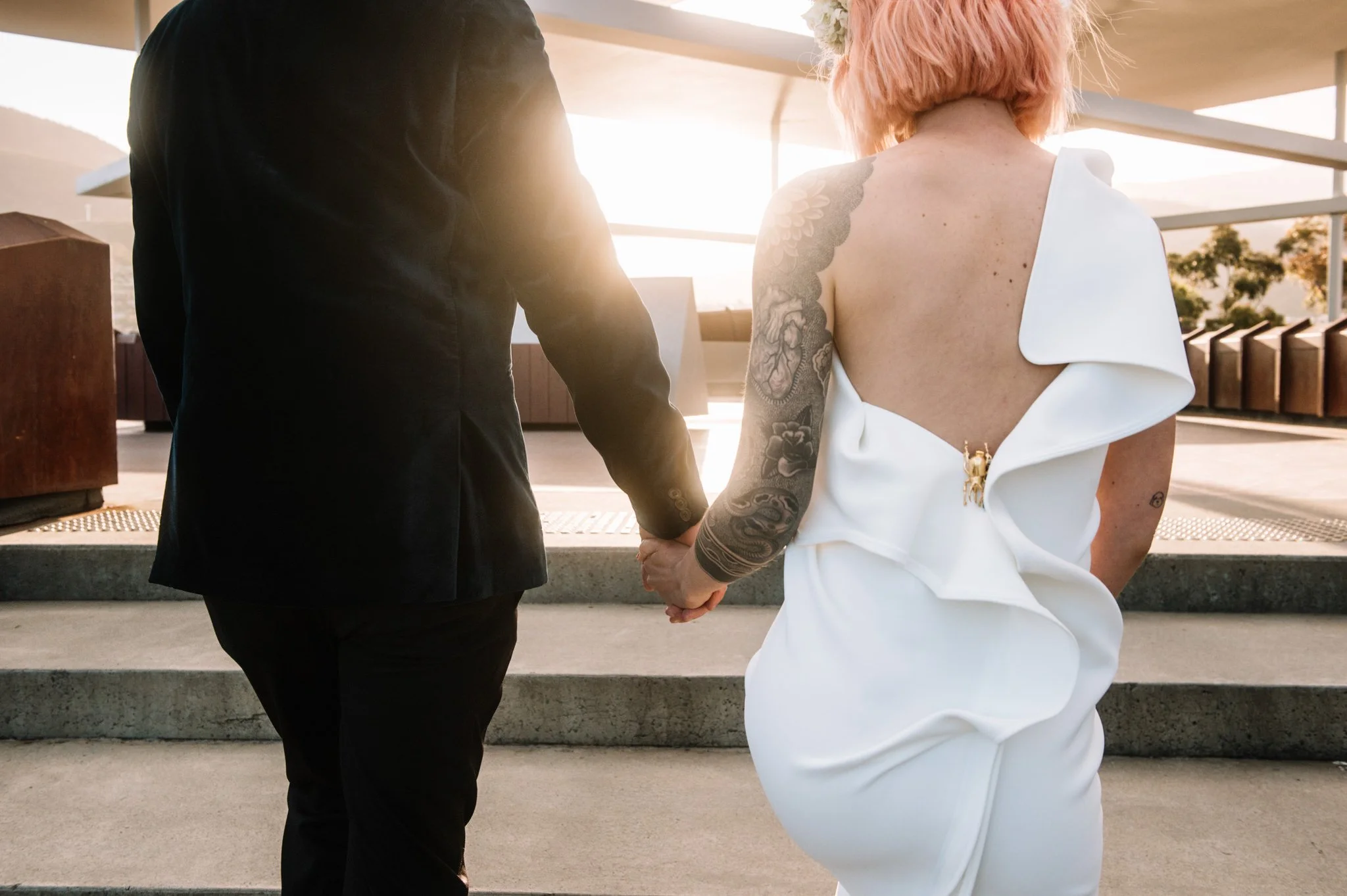 A couple holding hands during their wedding, with the bride wearing a white wedding dress with a ruffled sleeve and pink hair, and the groom in a black suit, standing outdoors on steps with sunlight shining behind them.