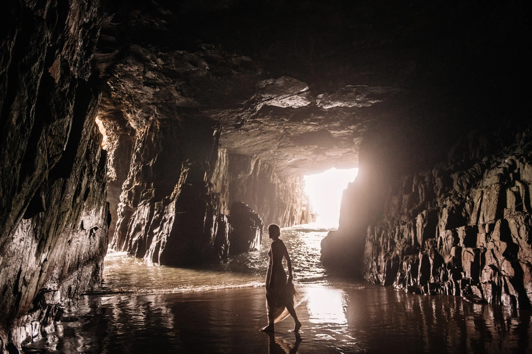 Inside a dark cave with textured rock walls and an opening to the bright outside, where two people are walking near the water's edge.