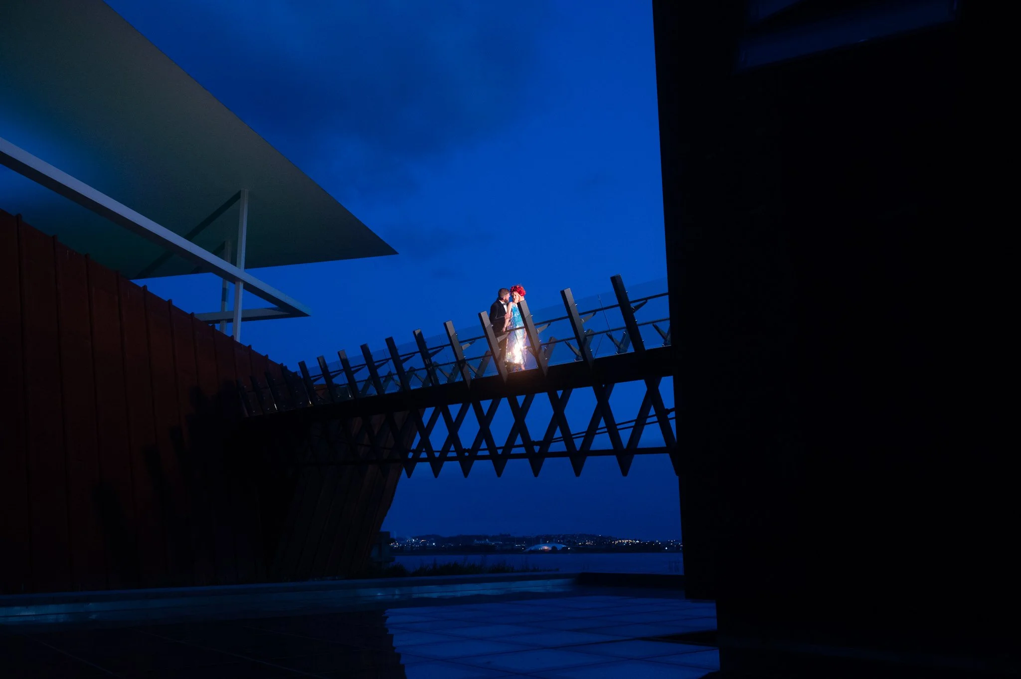 A romantic couple standing on a modern balcony at dusk, overlooking a body of water with city lights in the background.