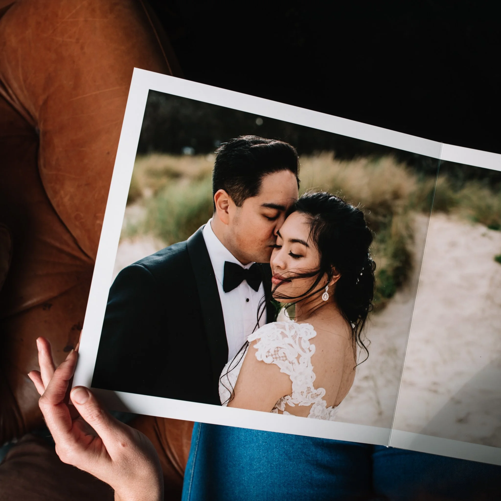 A person holding a framed photo of a couple, with the person in the photo dressed in a tuxedo and the woman in a white lace dress, appearing to be at an outdoor wedding.