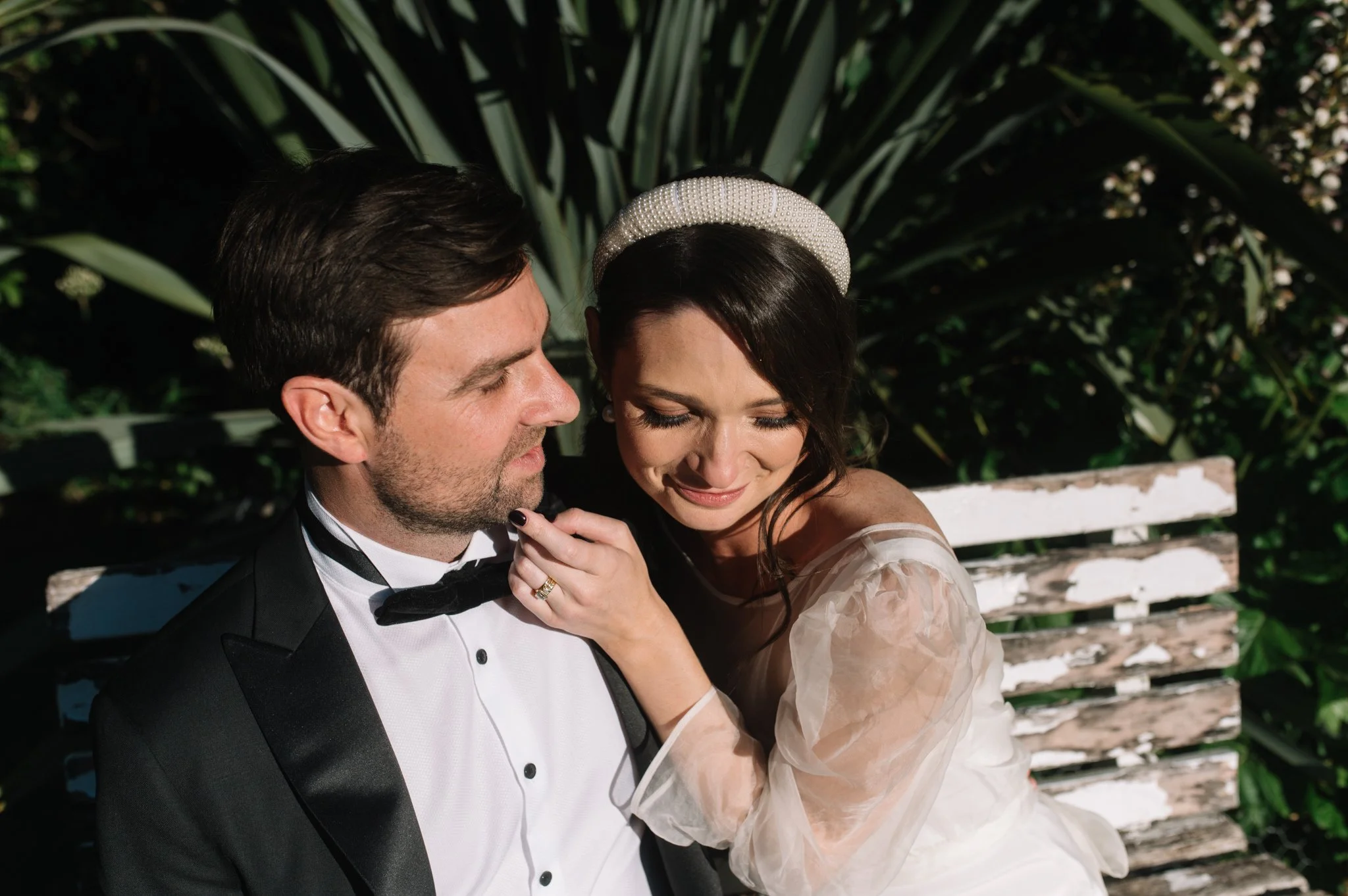 A bride and groom sit close together on a white bench outdoors, with the groom in a tuxedo and the bride wearing a pearl headband and a light-colored dress. They appear happy and intimate, surrounded by greenery.