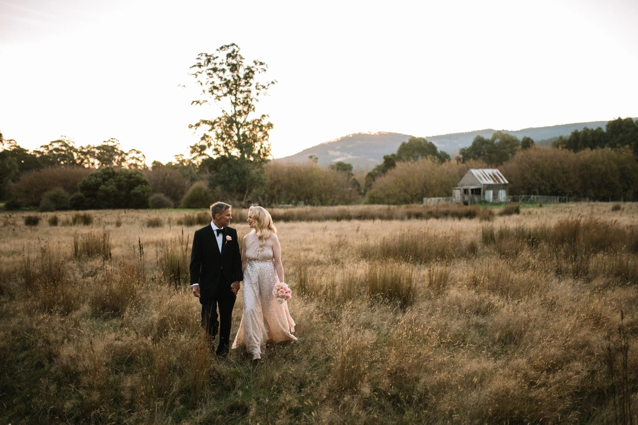 A bride and groom walking hand in hand across a grassy field during sunset, with trees and a small wooden shed in the background.