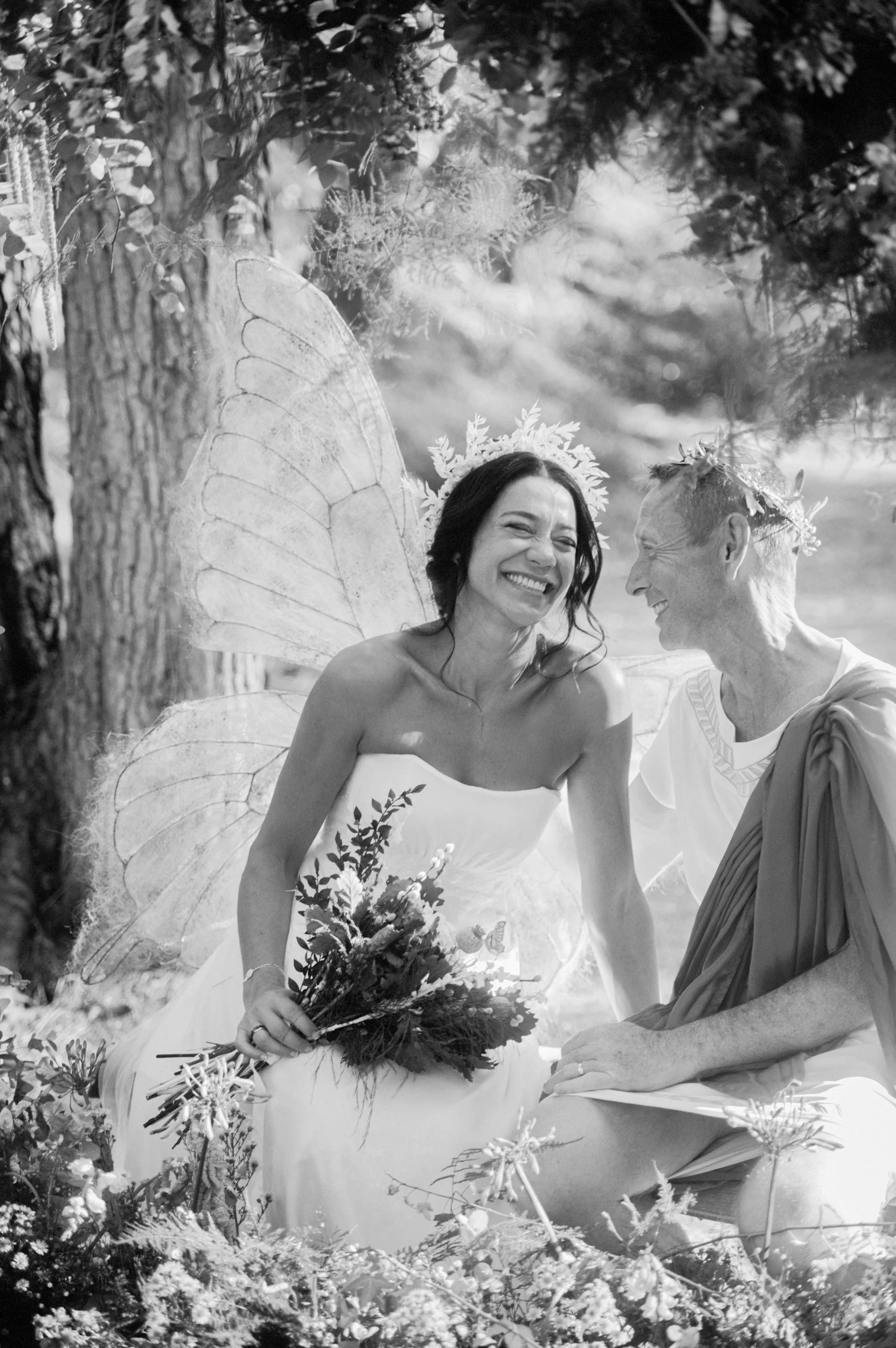 A black-and-white photo of a smiling woman dressed as a fairy with fairy wings and a floral crown, holding a bouquet, sitting next to a man wearing a toga and a floral crown, in an outdoor garden setting.