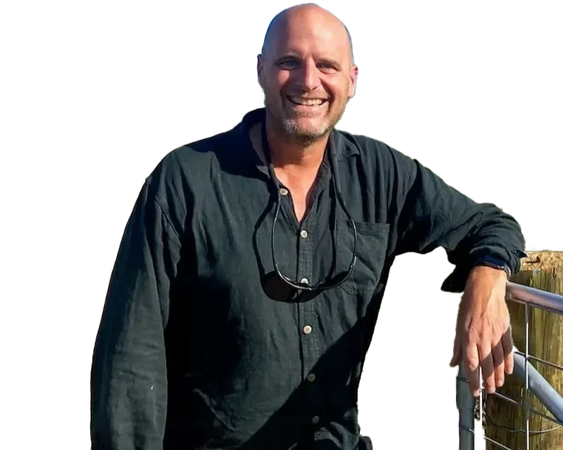 A smiling bald man with a beard, wearing a dark button-up shirt with the sleeves rolled up, standing outdoors with a railing, during daytime.