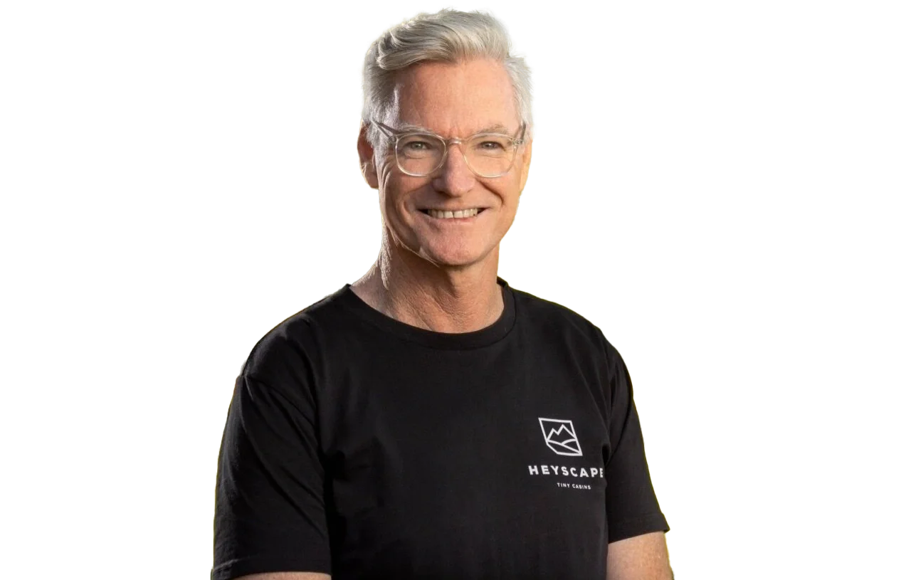 A smiling older man with gray hair and glasses wearing a black T-shirt with a logo that says "Heyscap".