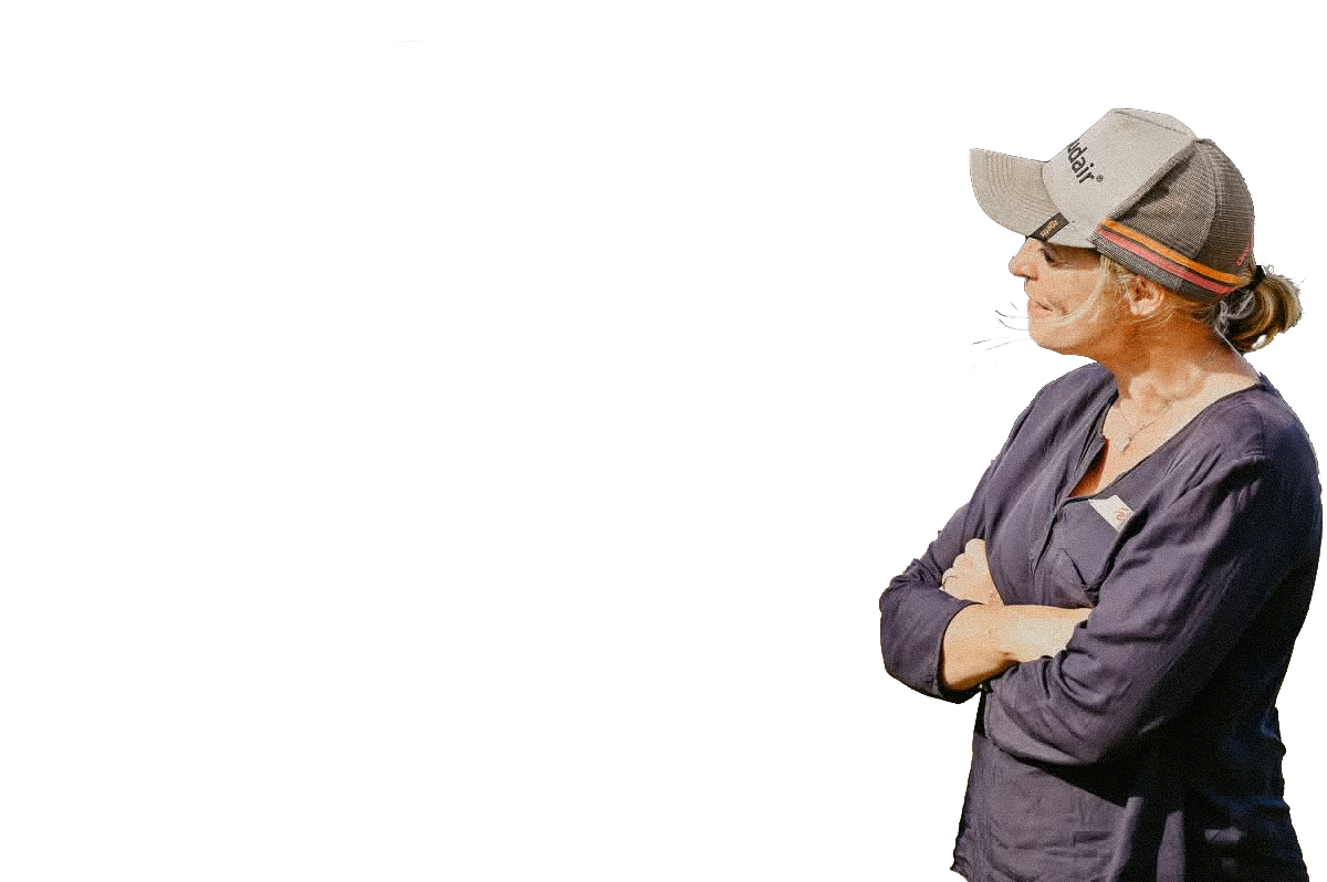 A farming woman with her arms crossed wearing a gray baseball cap with orange and black stripes, a dark purple long-sleeved shirt, and glasses, looking to her left.