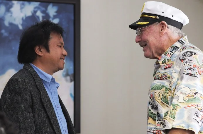 Two men smiling and facing each other, one in a dark blazer and blue shirt, the other in a colorful tropical shirt and a captain's hat, indoors with a world map on the wall.