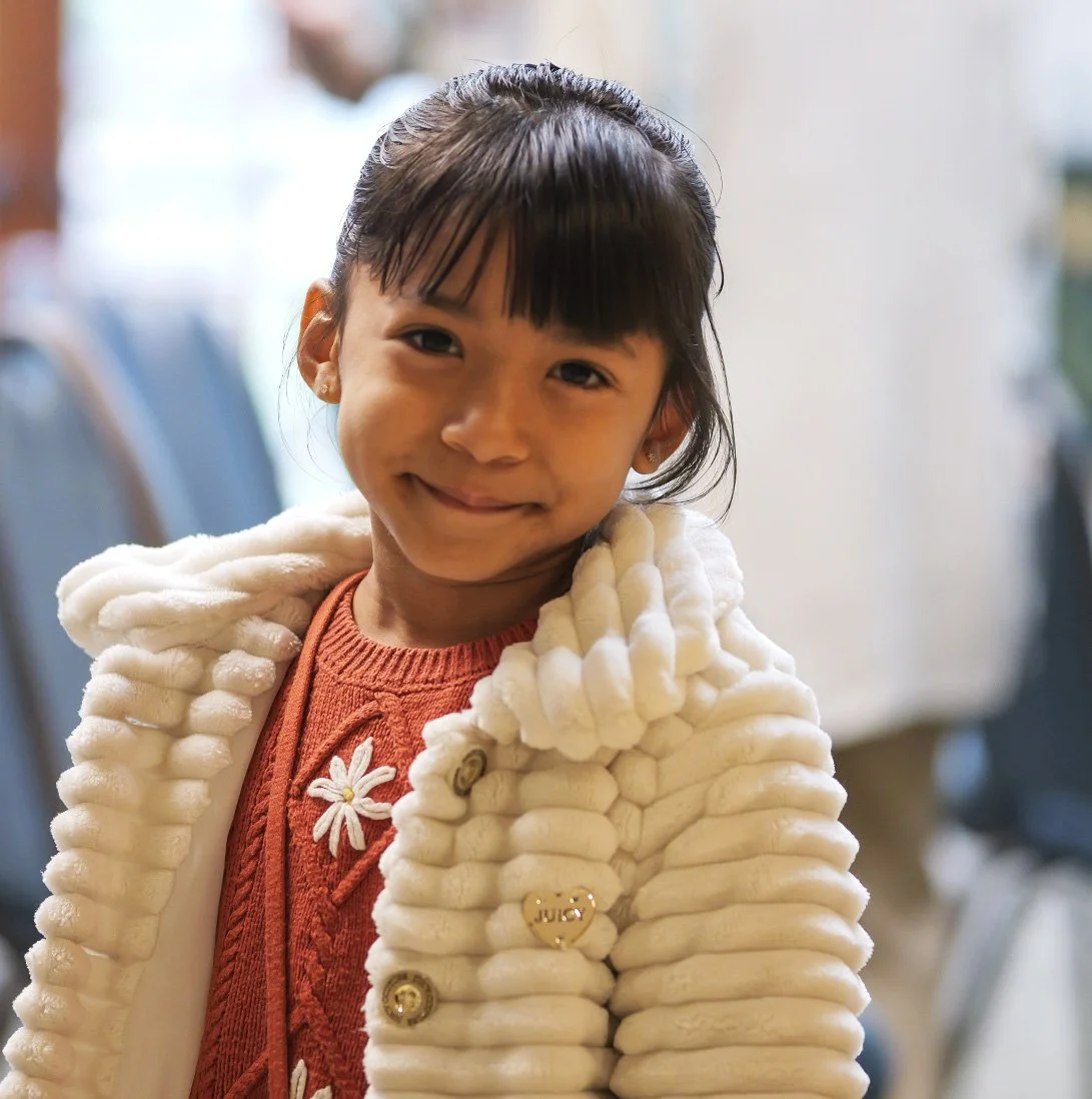 A young girl with dark hair tied back, smiling, wearing a white fuzzy coat over a red sweater with flower embroidery, indoors with blurred background.