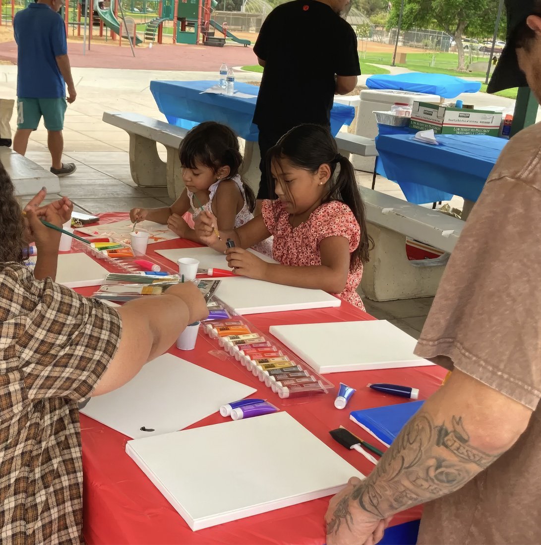 Children painting on blank canvases at an outdoor art activity table.