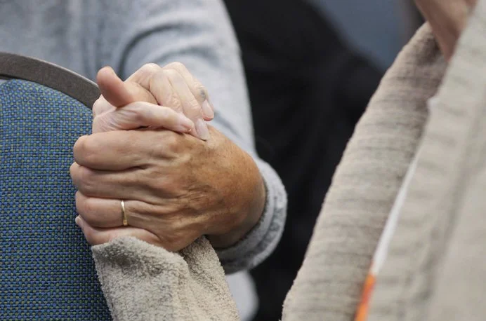 Two elderly people holding hands, one with a wedding ring, sitting in chairs.