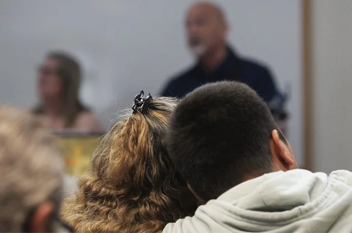 Two people sitting close together, viewed from behind, listening to a speaker at a meeting or classroom.