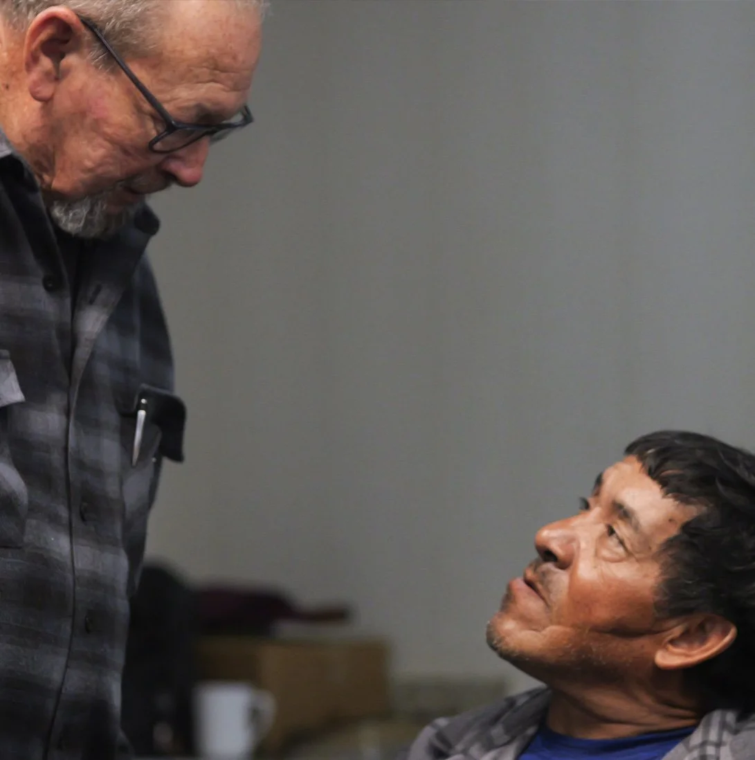 An elderly man and a middle-aged man are having a conversation indoors, with the elderly man standing and the middle-aged man sitting, looking up at the standing man.