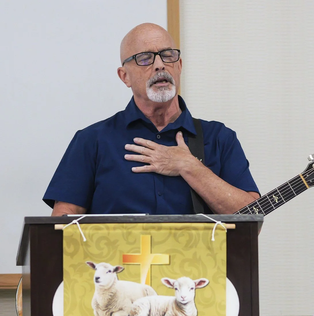 An elderly man with glasses and a white beard, wearing a navy blue shirt, stands behind a podium with a religious banner featuring sheep and a cross. He is holding his chest and has his eyes closed with an expression of emotion.