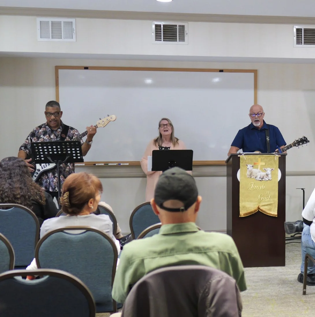 People singing and playing guitars in front of an audience in a room with a whiteboard, religious banners, and ceiling vents.