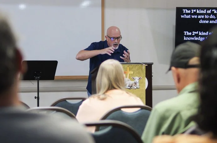 A speaker, an older man with glasses, stands behind a podium with a lamb and cross symbol, gesturing with his hands during a presentation in a classroom or conference room. Audience members are seated facing him, with a large screen displaying text in the background.